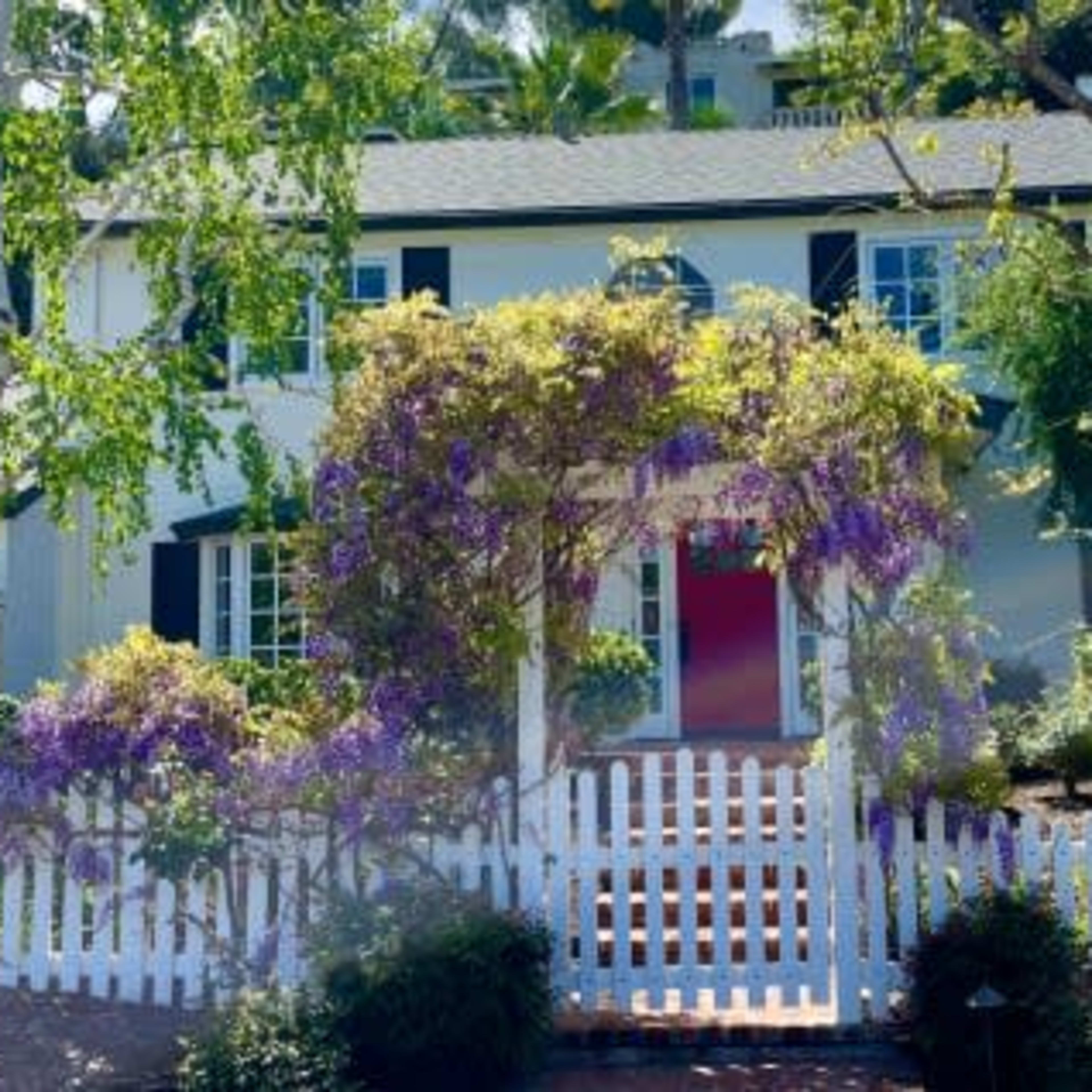 A two-story house with white walls, black shutters, and a vibrant entrance framed by purple flowers and a white picket fence.