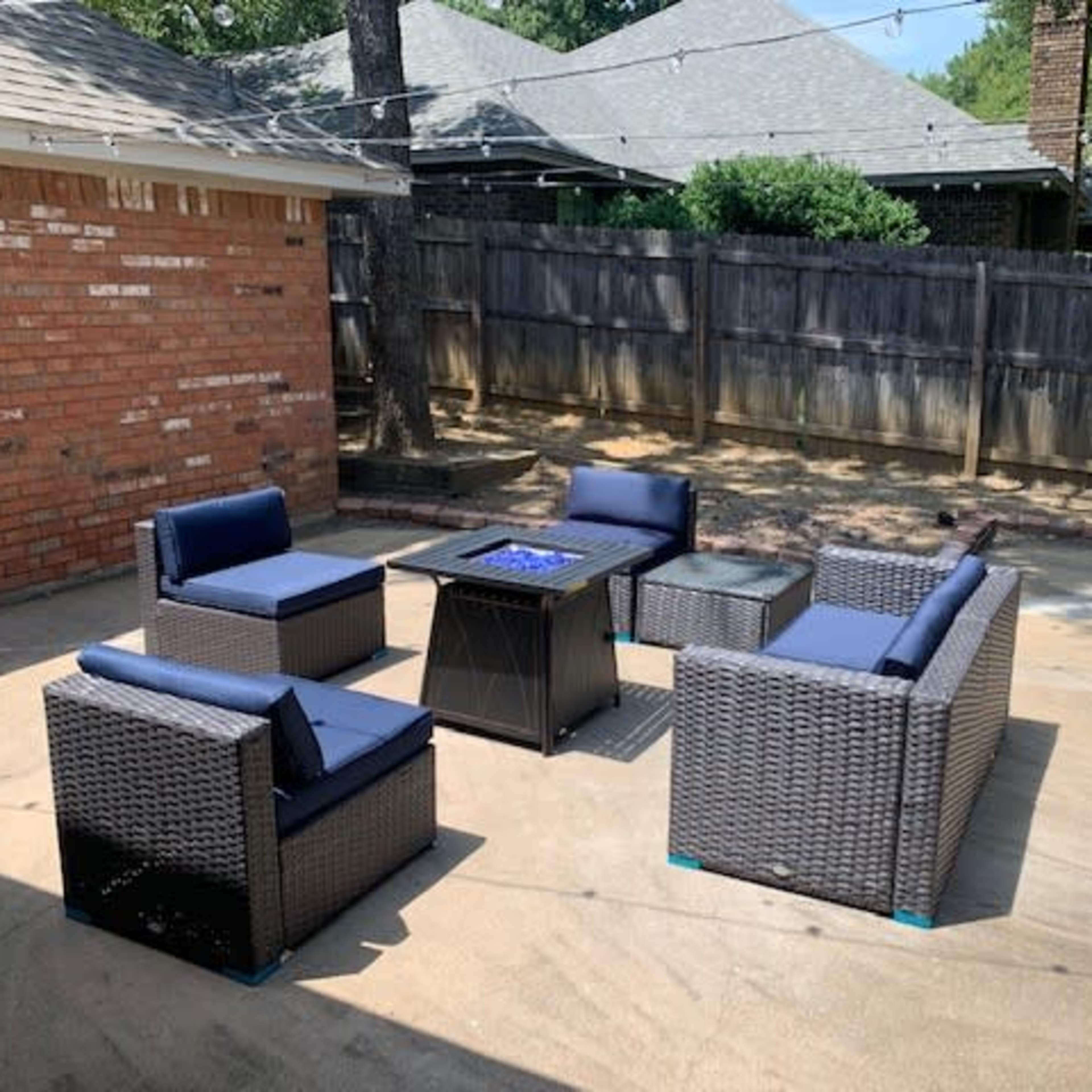 A patio area features four wicker chairs with blue cushions surrounding a square fire pit on a concrete surface.