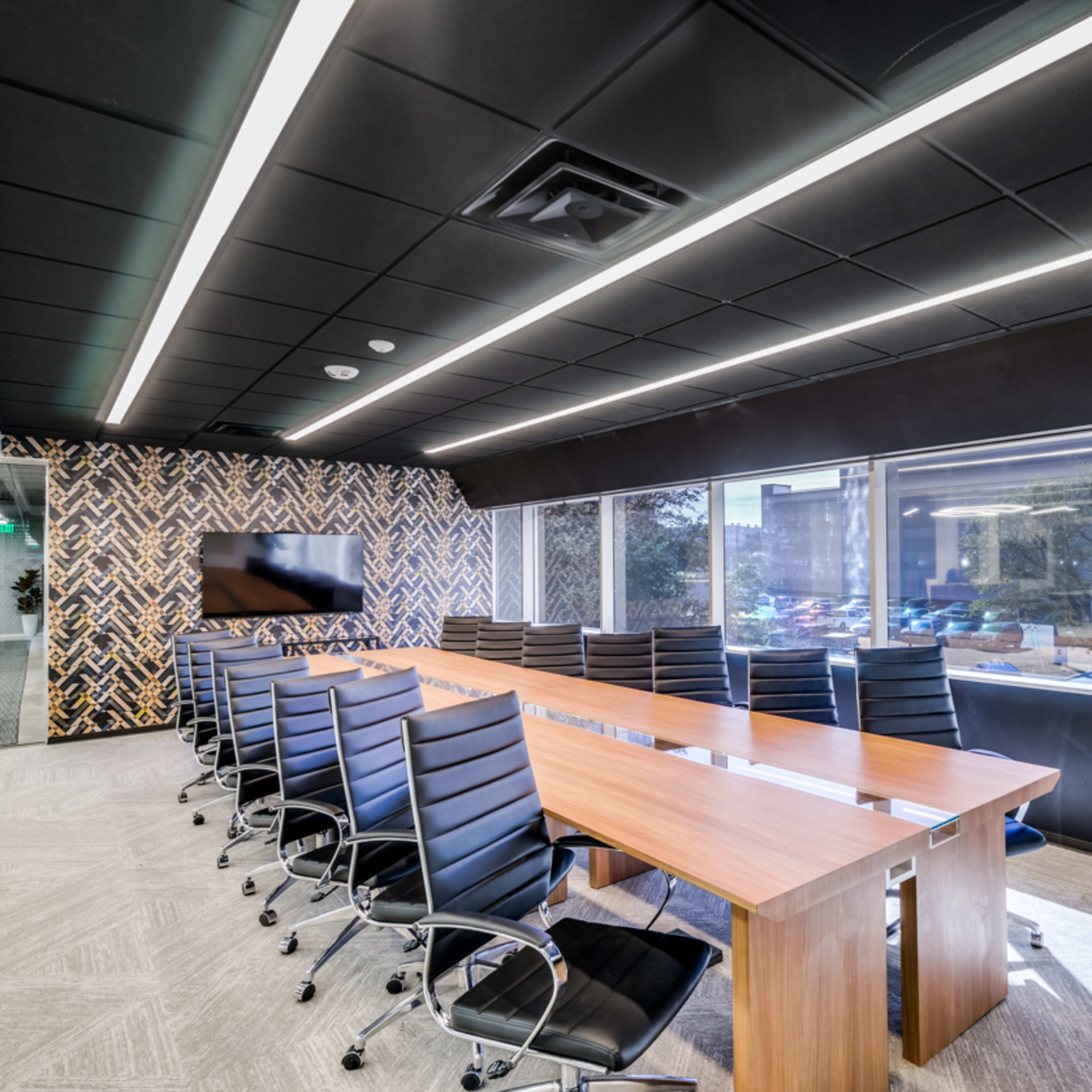 A modern conference room features a large wooden table surrounded by black ergonomic chairs, with large windows providing natural light and views of the outside.