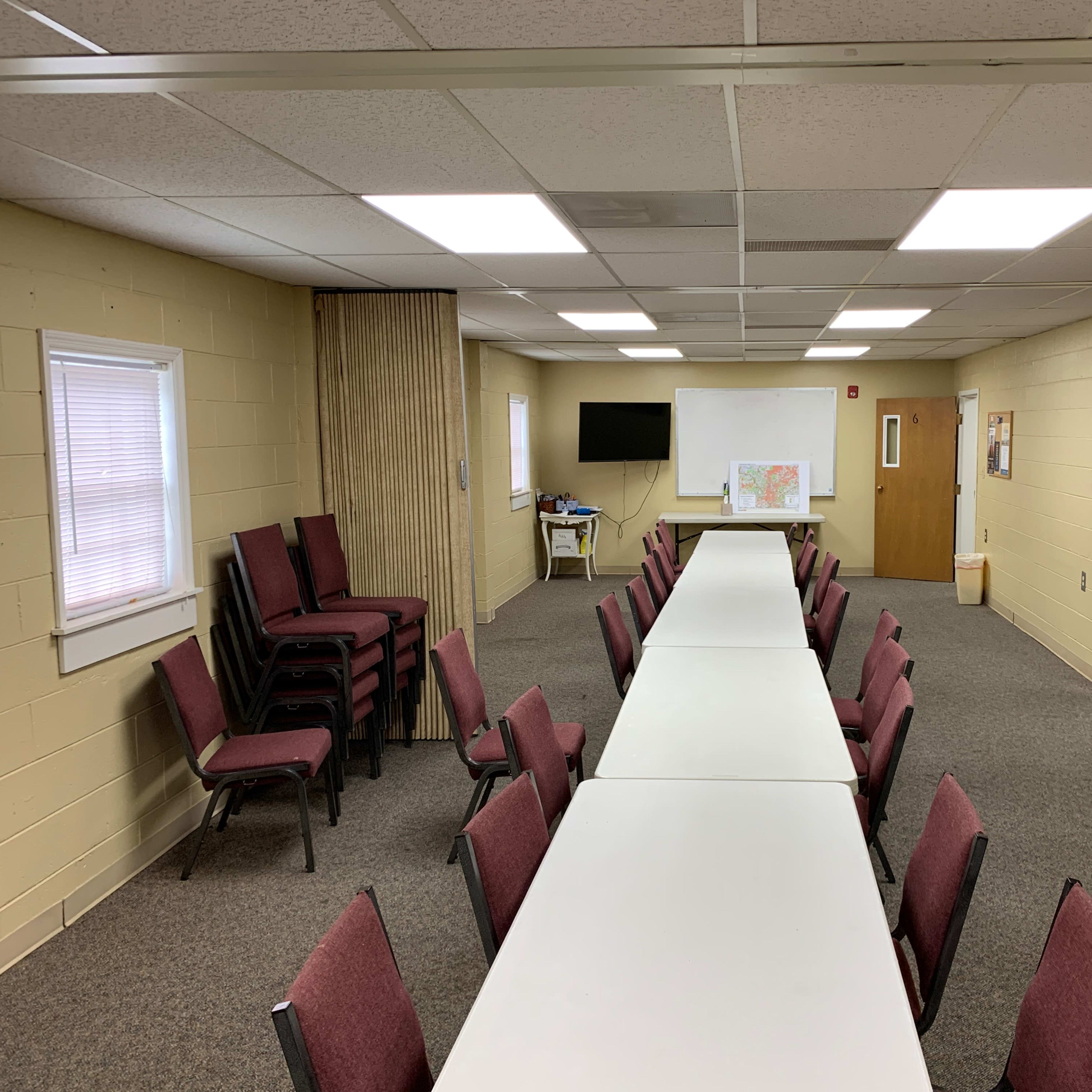 A long, rectangular table with chairs on either side is set up in a spacious room featuring beige walls, multiple light fixtures, and two doors.