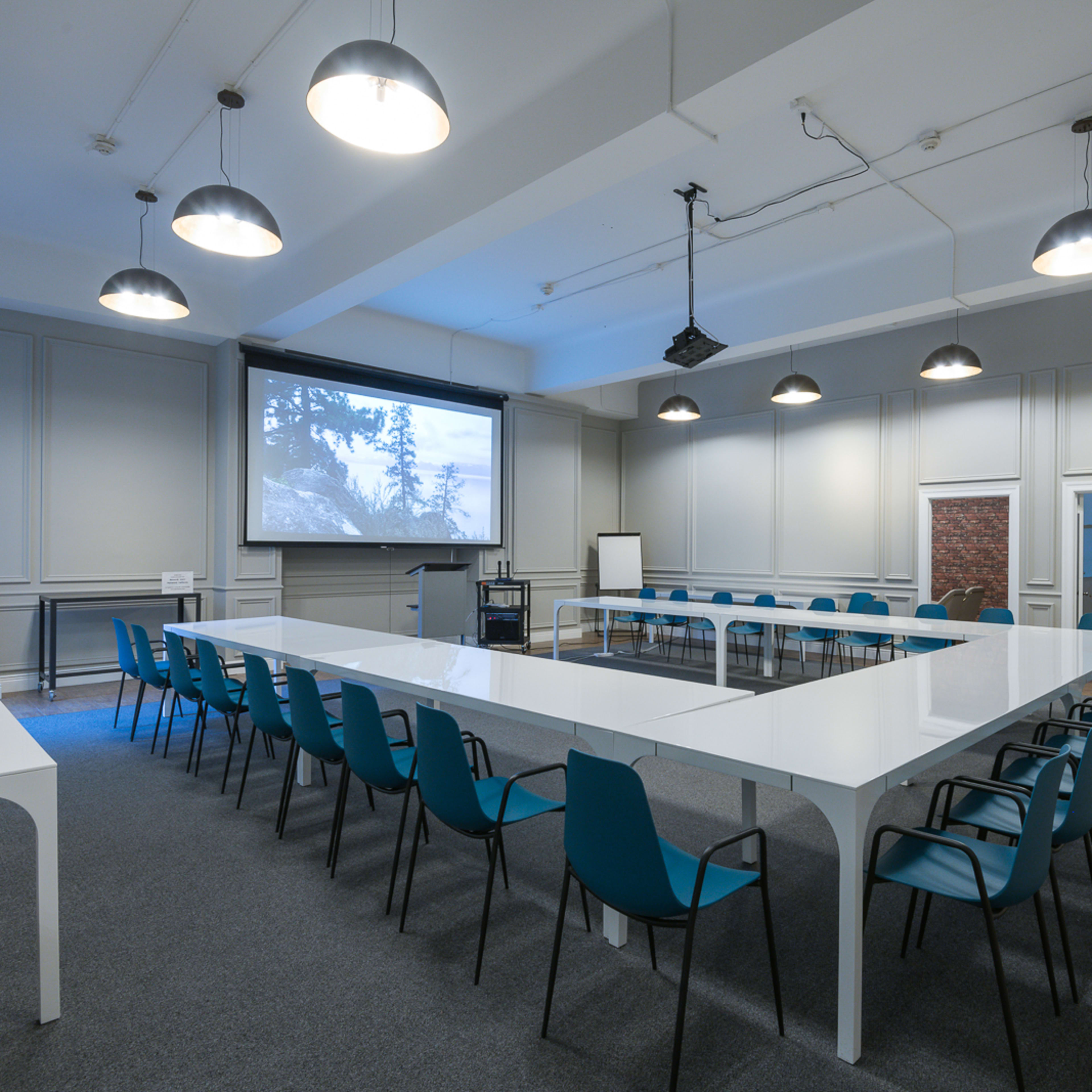 A modern meeting room featuring a long white table surrounded by turquoise chairs, with a projector screen at one end and various seating arrangements.