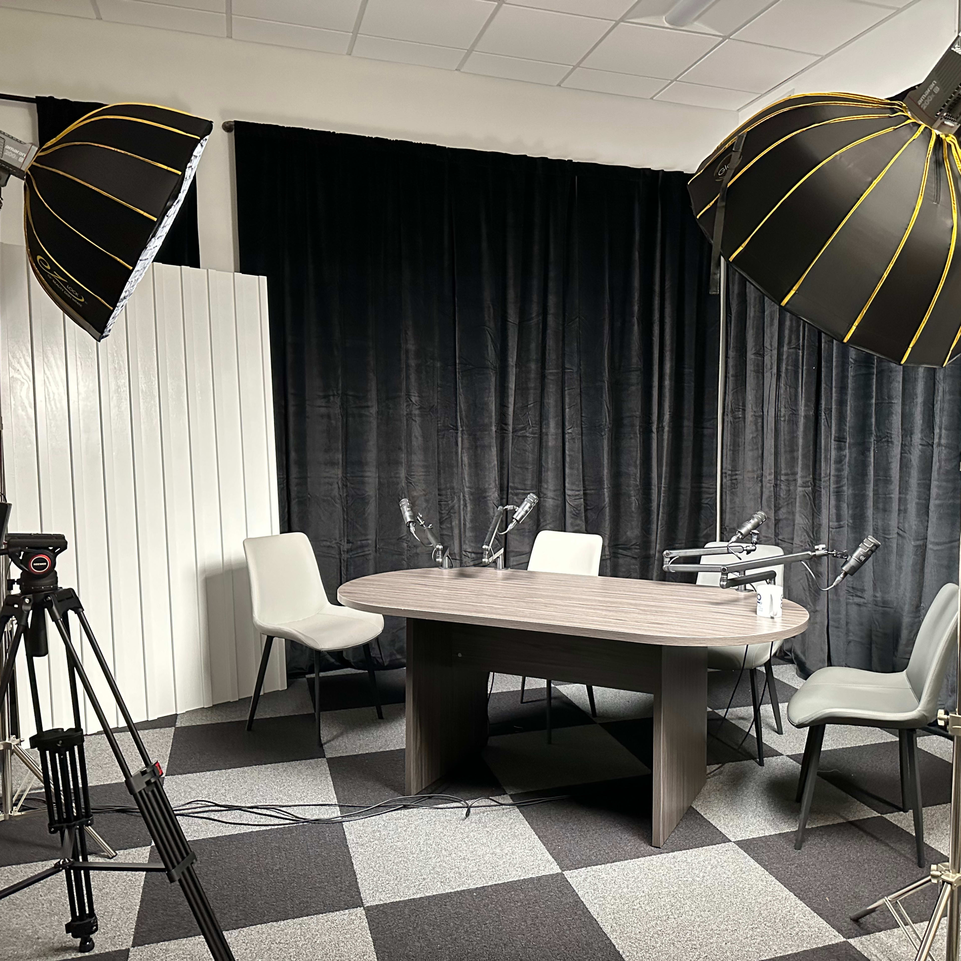 A recording studio setup with a round table, four chairs, and two large light fixtures, all surrounded by black curtains.