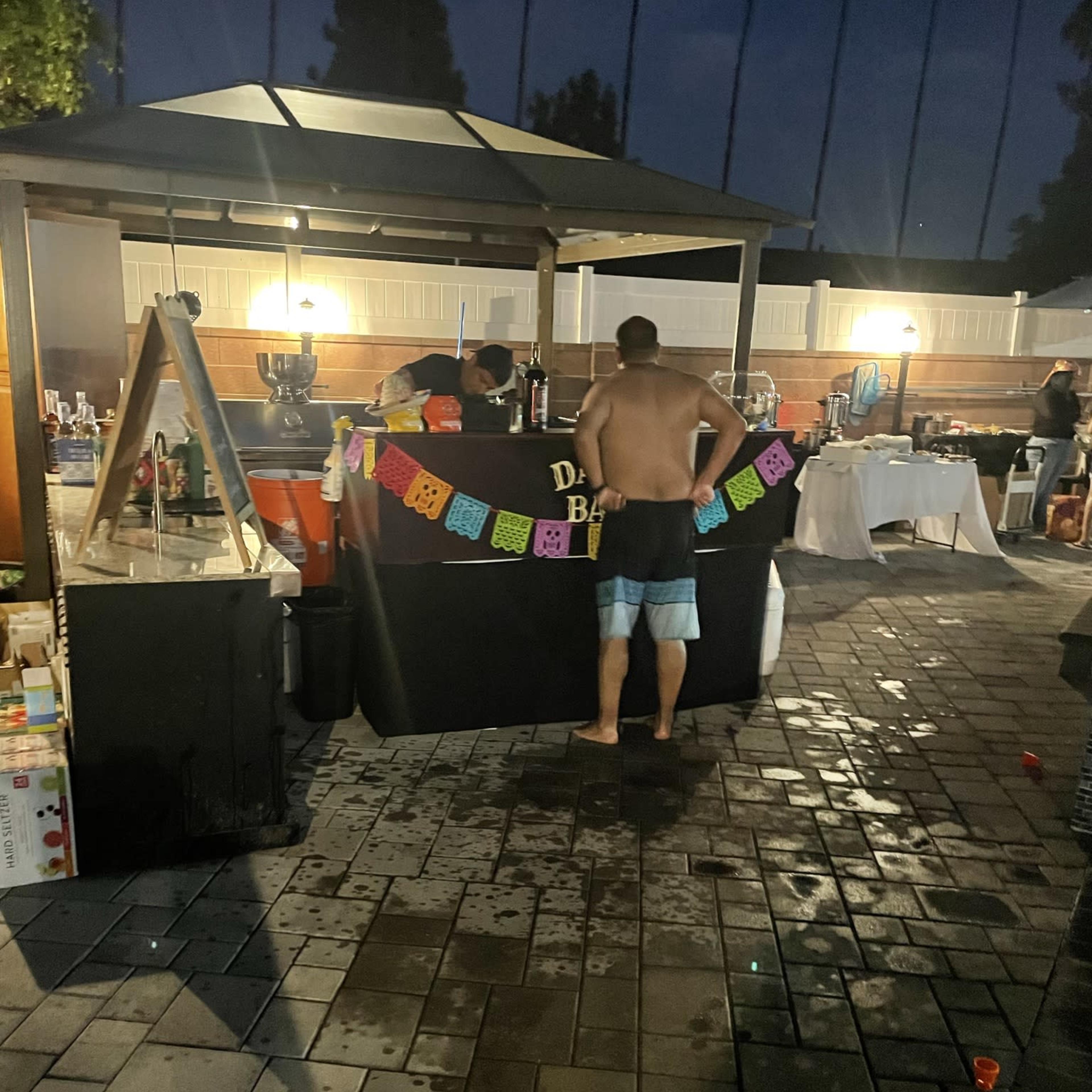 A shirtless man stands in front of a decorated bar counter under a gazebo at a nighttime outdoor event.