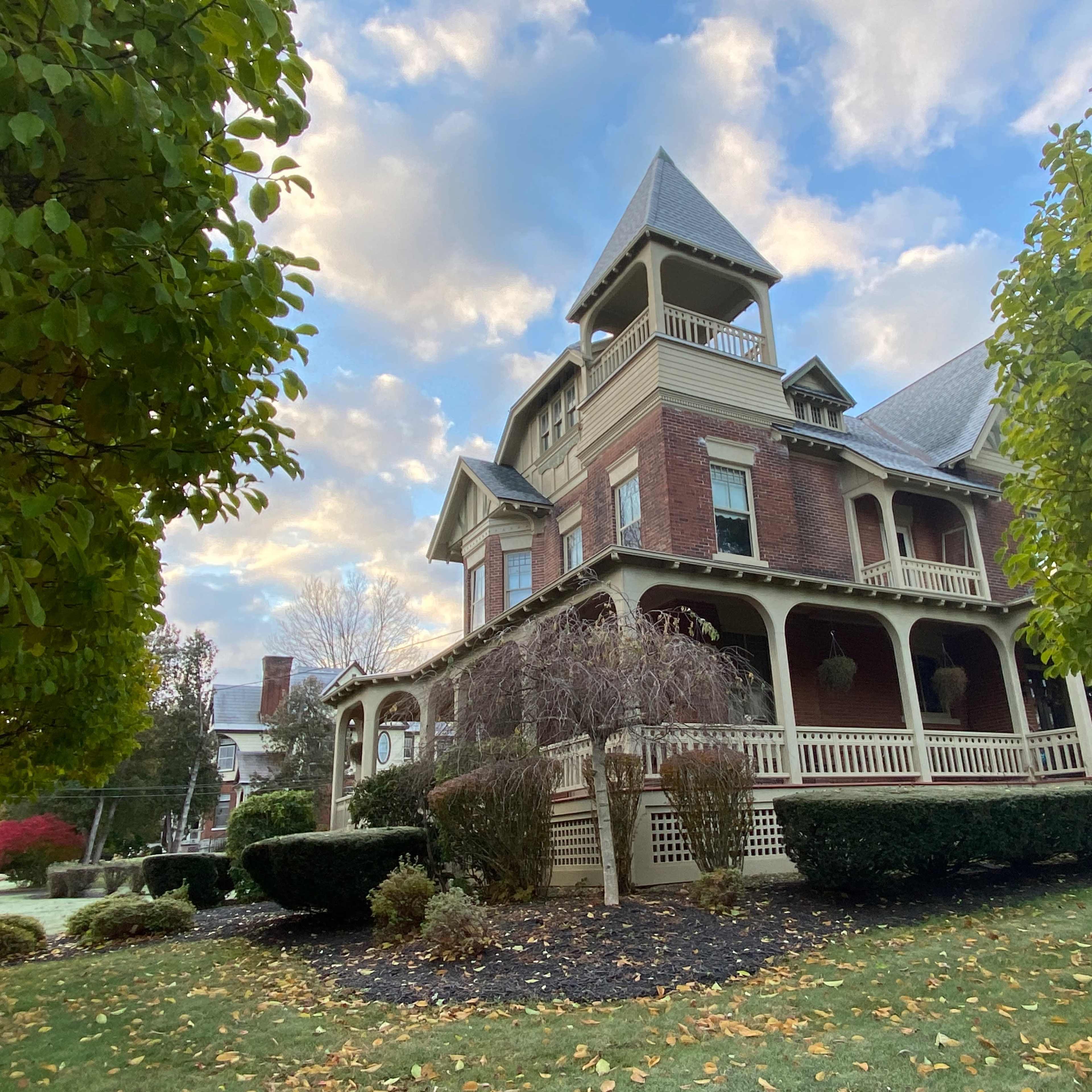 A large, historic brick house with a turret and a wraparound porch is surrounded by well-manicured lawns and trees.