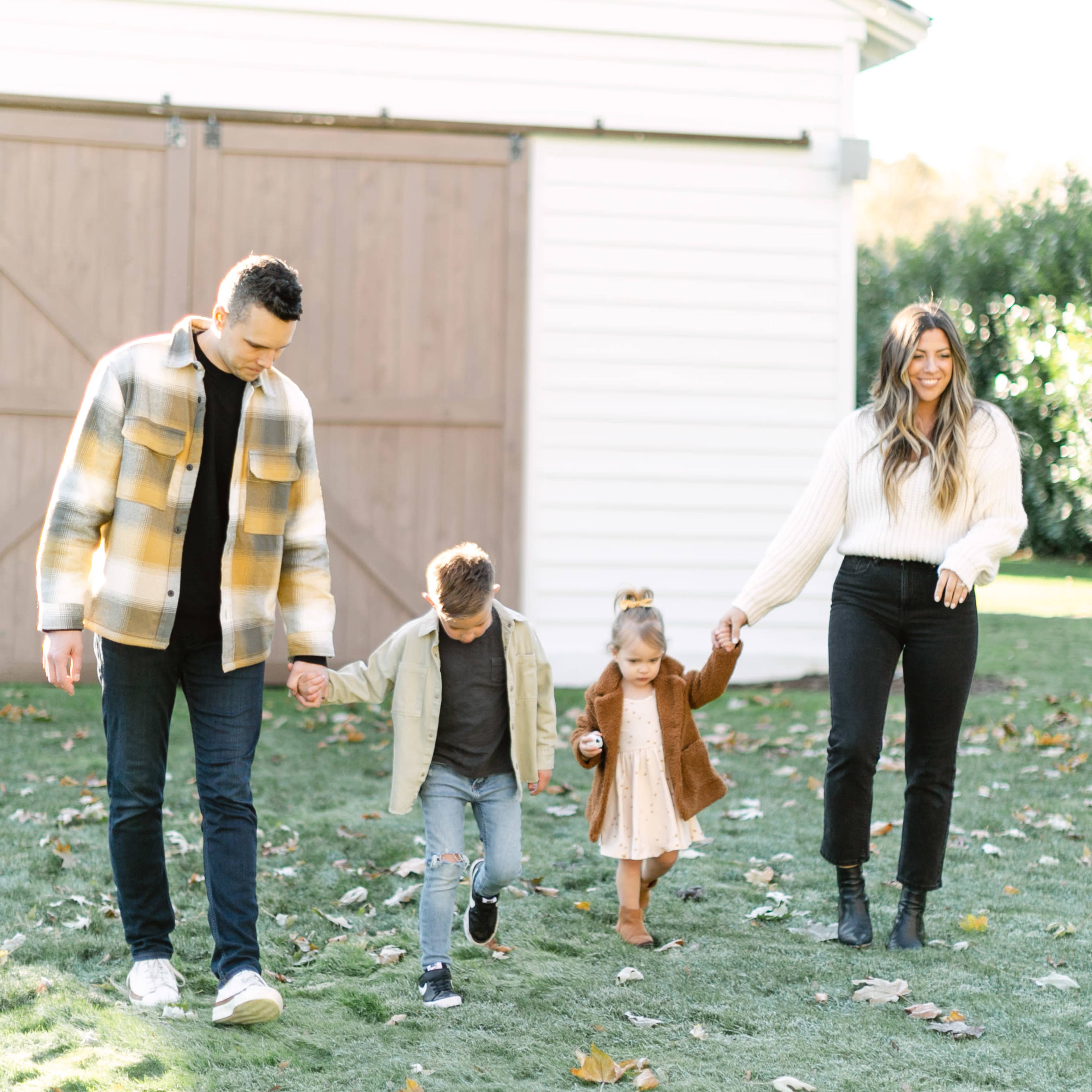 A family of four walks hand in hand on a grassy area near a white barn.