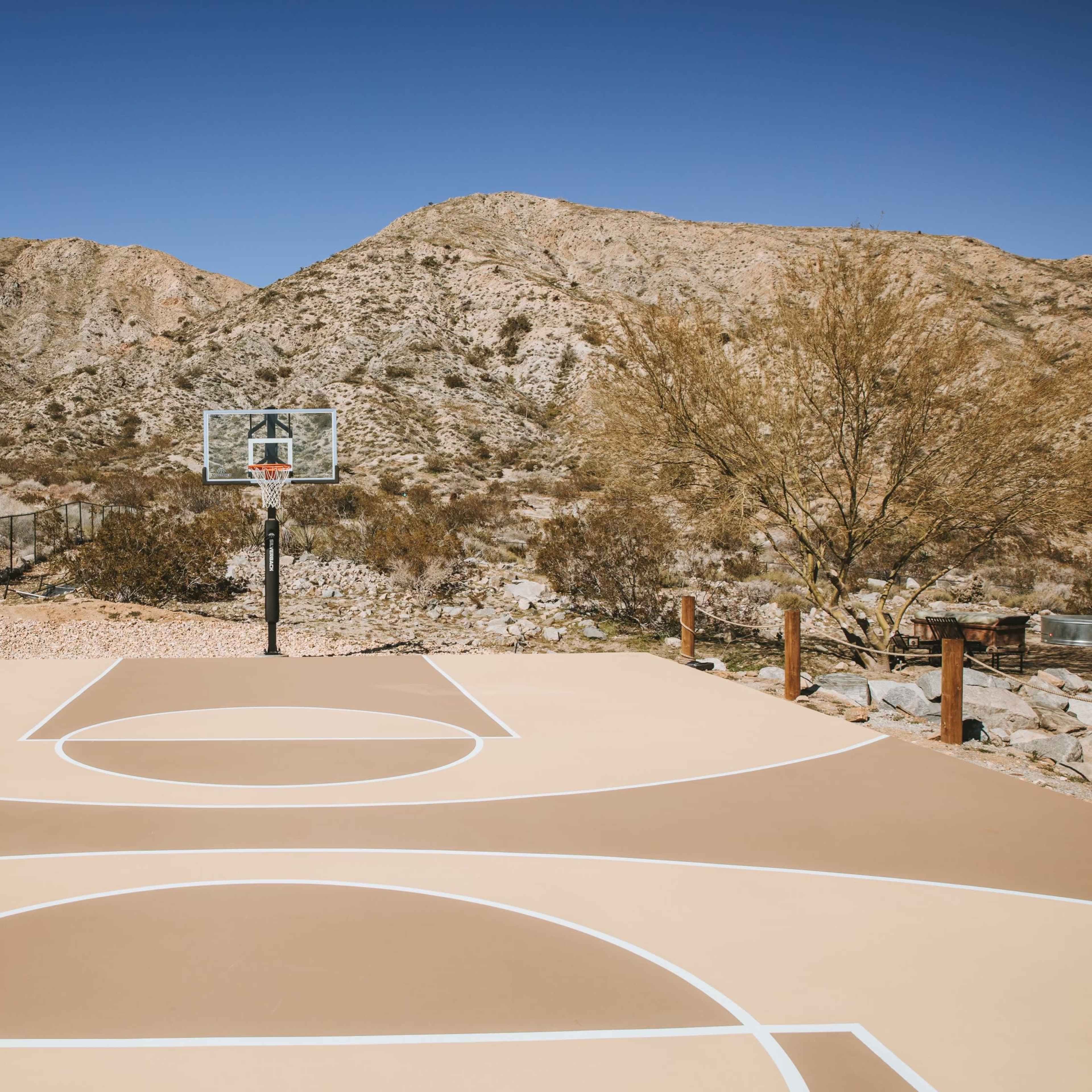 A basketball court with a hoop stands in a desert landscape framed by mountains under a clear blue sky.