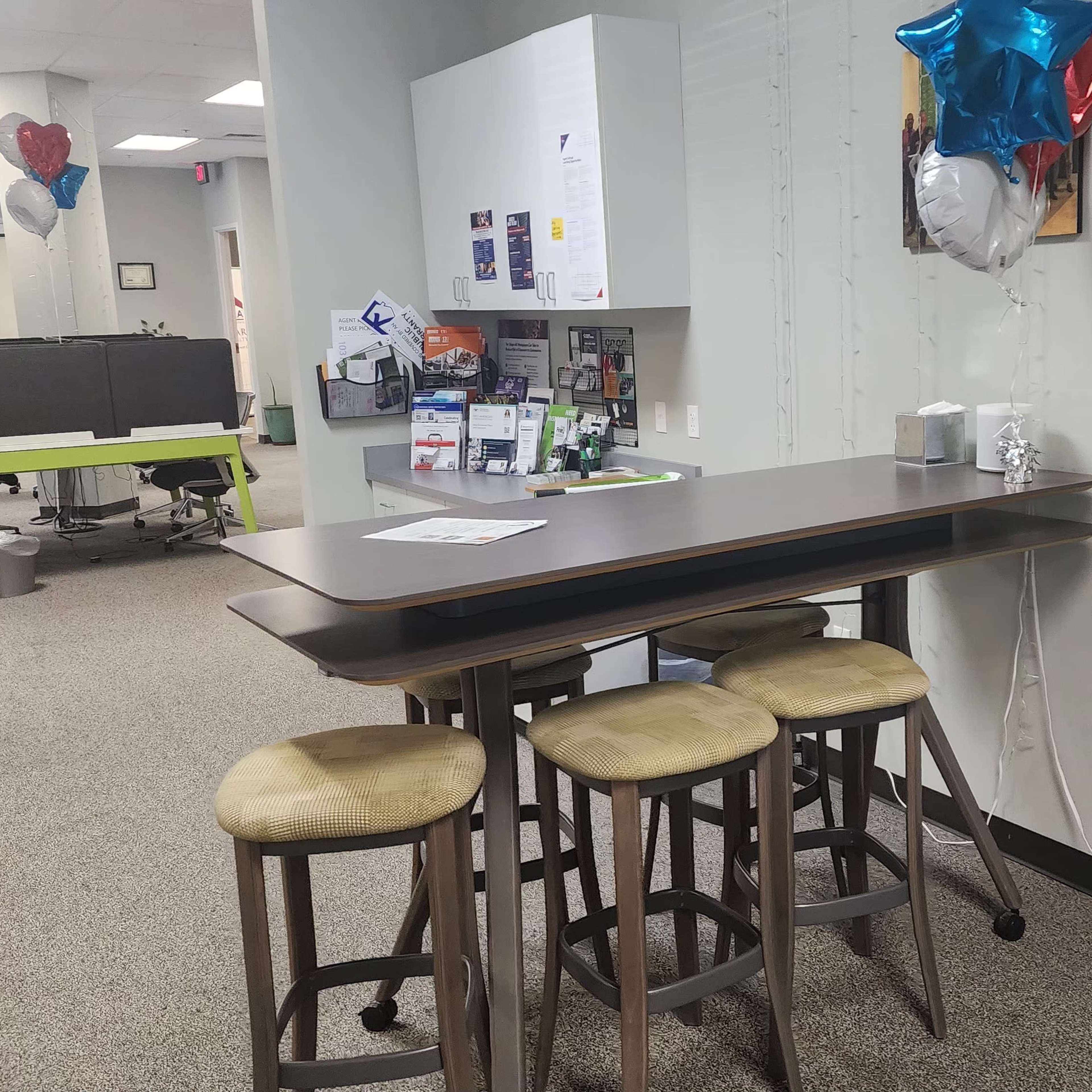 A small kitchenette area with a high table and stools, decorated with balloons, in an office environment.