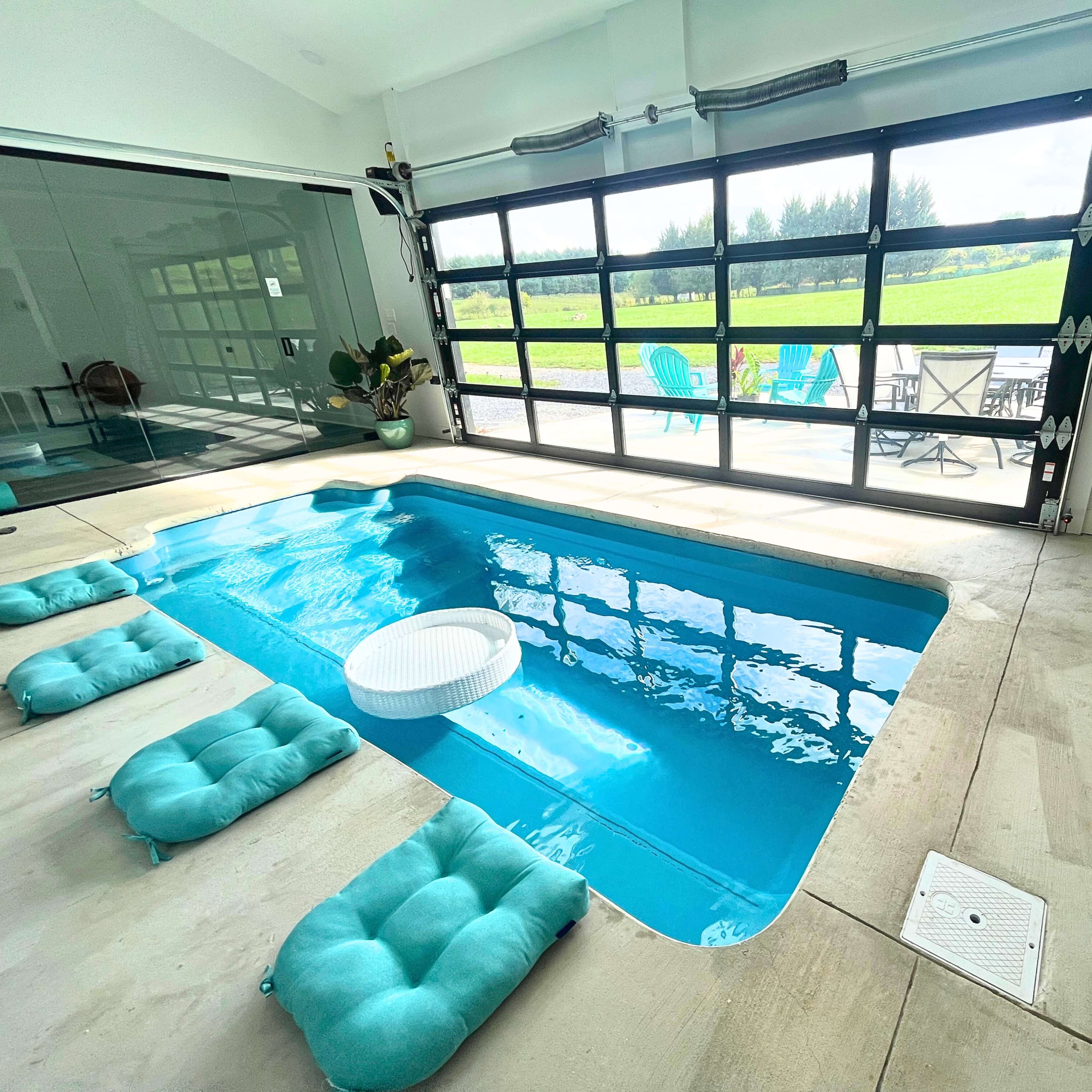 A modern indoor pool area with a glass door opening to a green outdoor space, featuring seating on both sides of the pool.