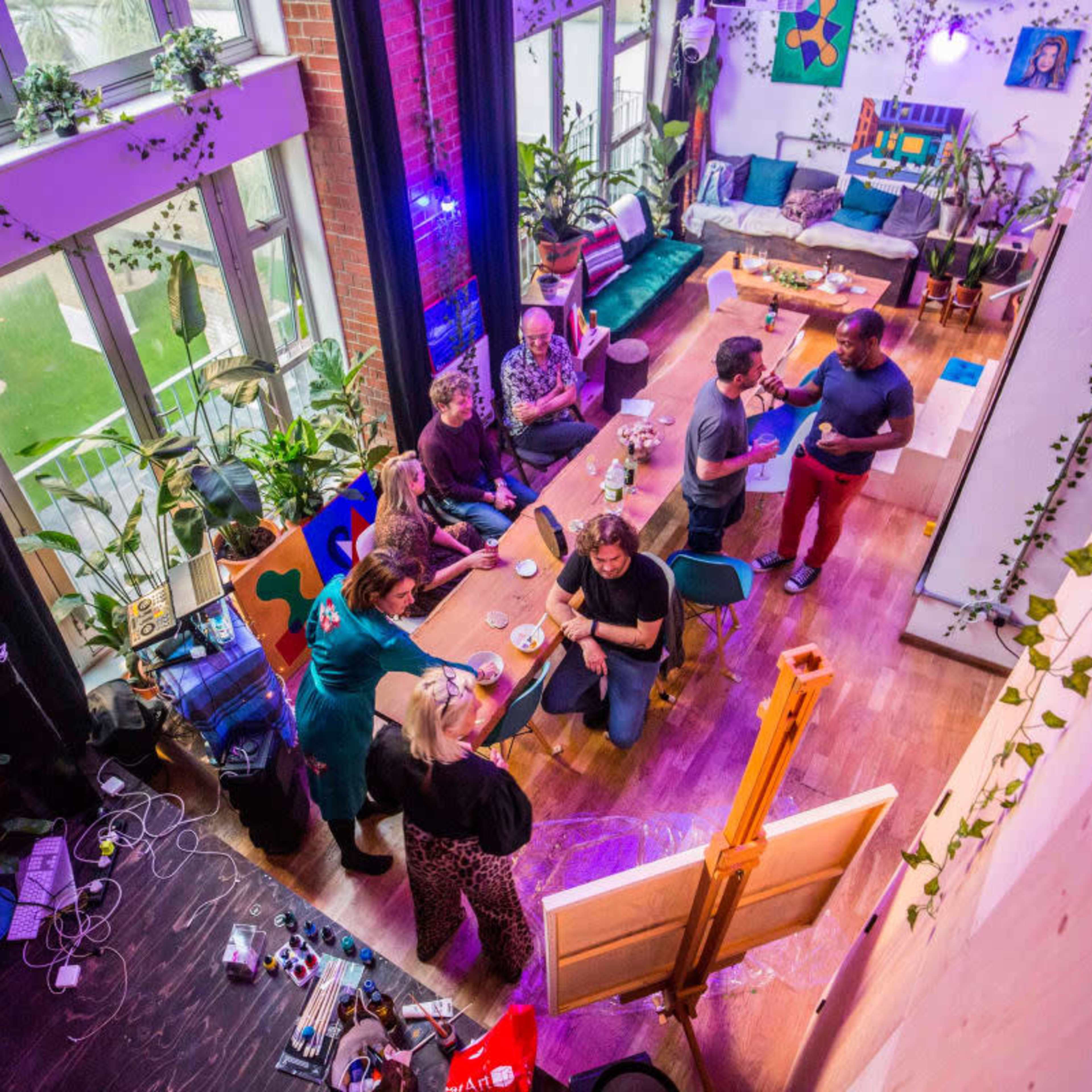 A group of people gather around a table in a colorful, plant-filled studio space while an artist works on a canvas nearby.