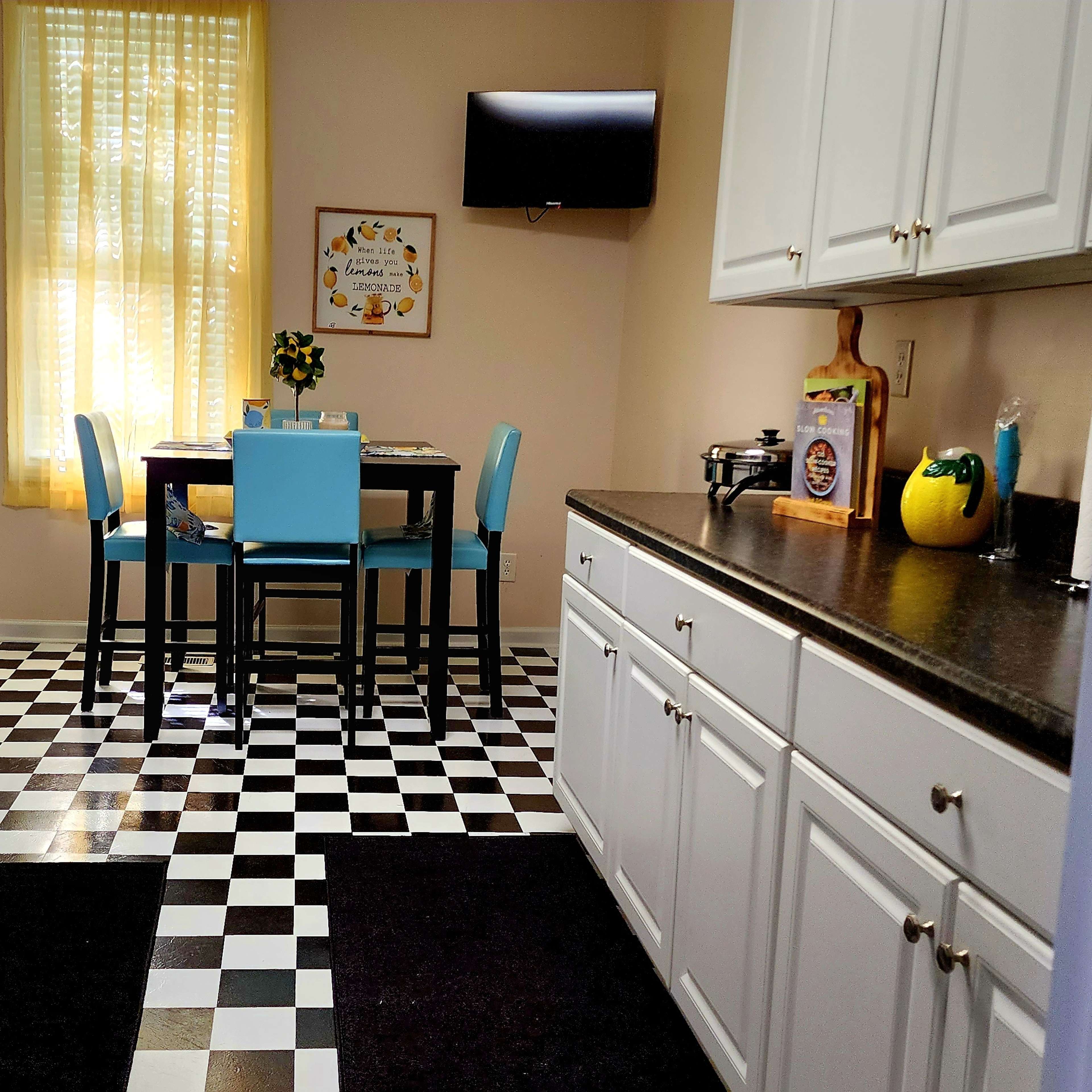 A tidy kitchen features white cabinets, black and white checkerboard flooring, a dining table with blue chairs, and a black refrigerator.