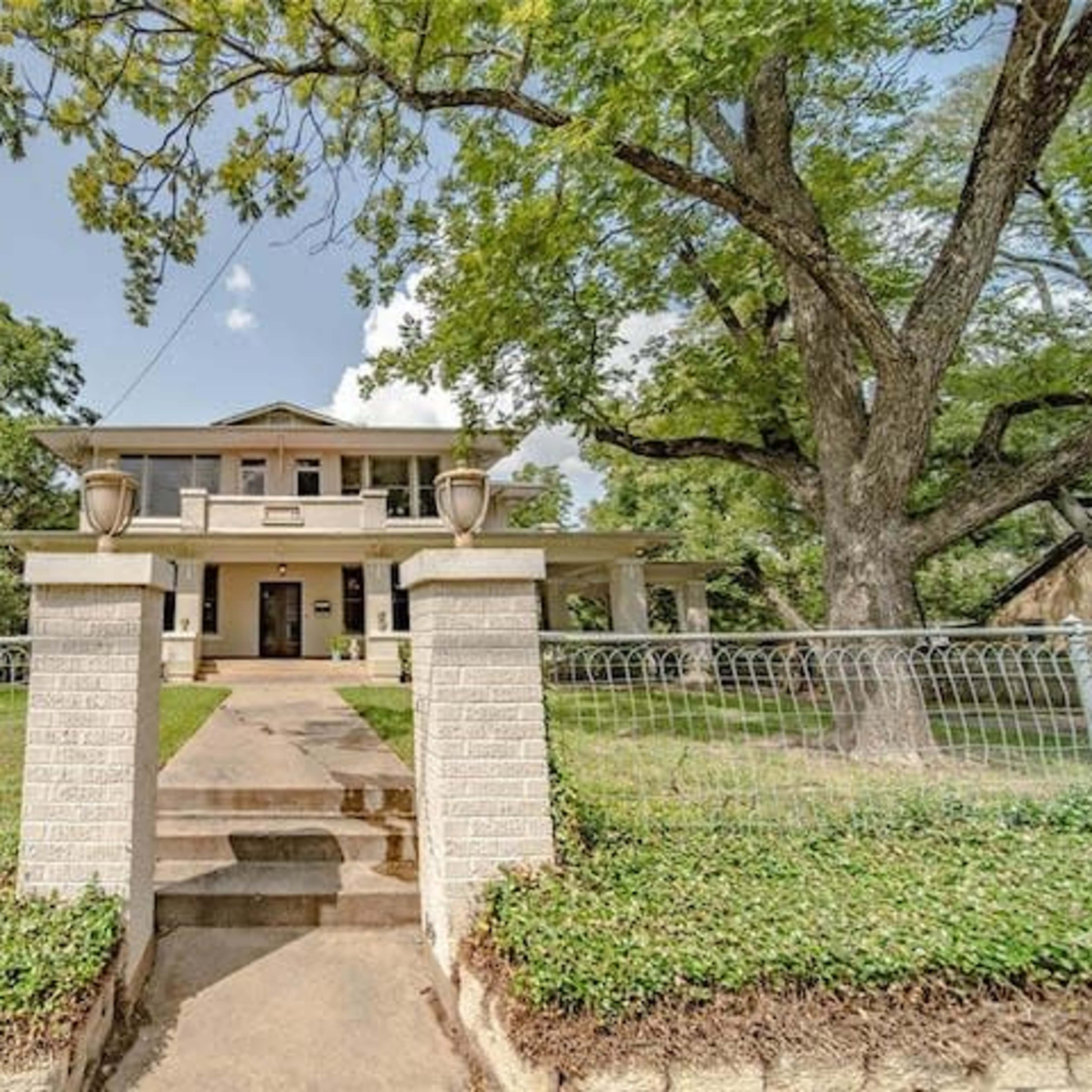 A two-story house with a large front porch is surrounded by trees and a decorative fence, featuring a pathway leading up to the entrance.