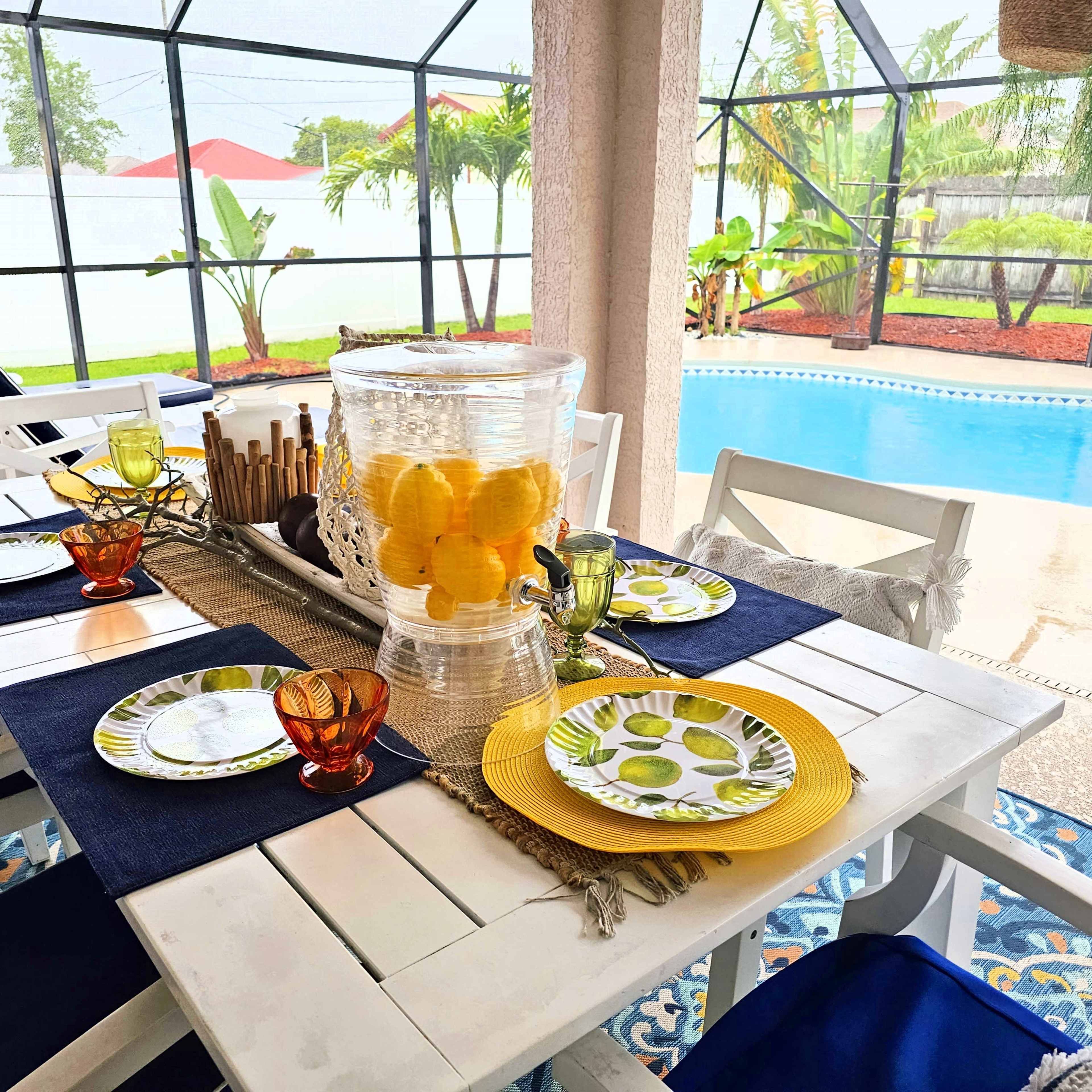 A dining table is set outdoors with colorful plates and glasses, alongside a large vase filled with lemons, beside a swimming pool surrounded by greenery.