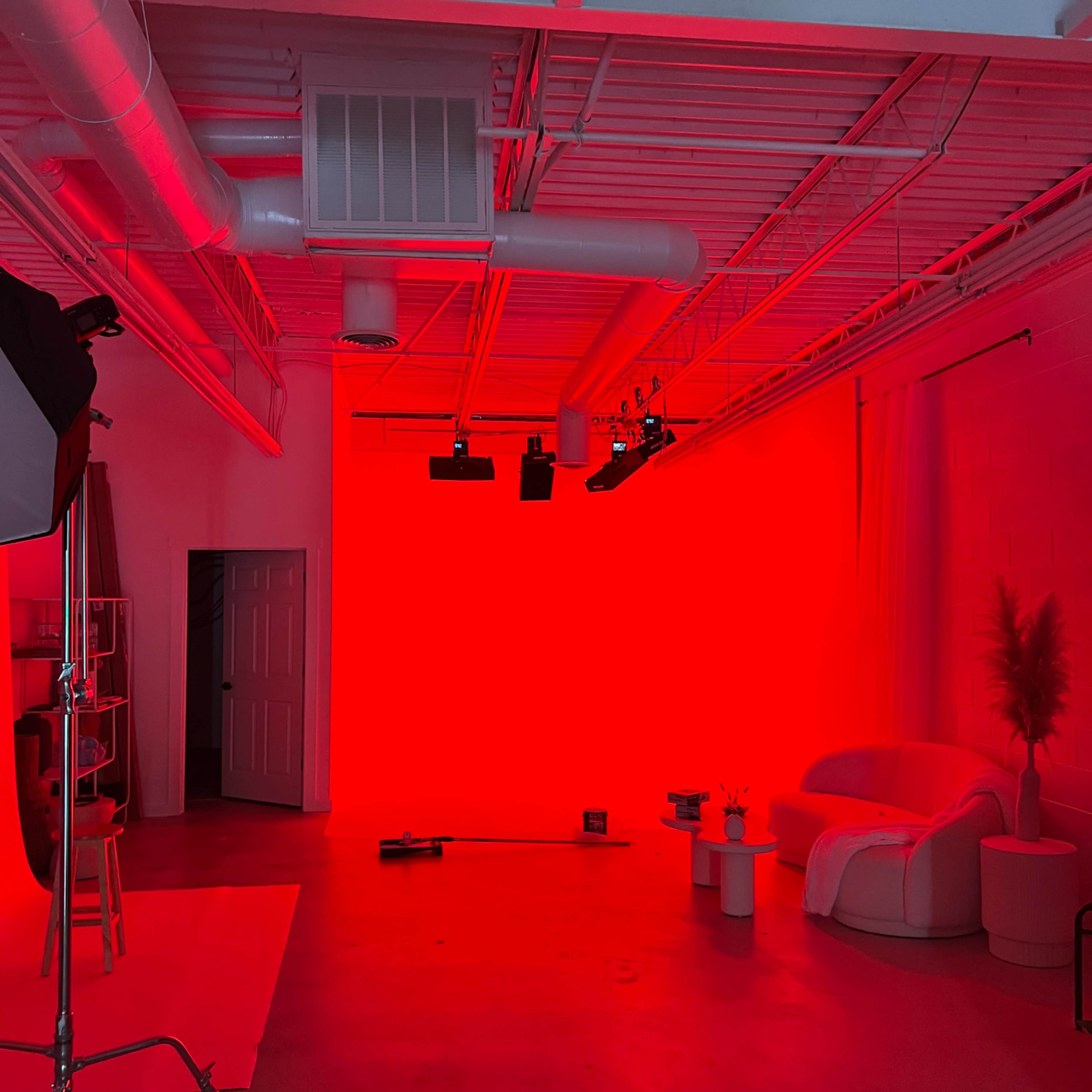 A studio space with a red backdrop, a white couch, a plant, and various lighting equipment.