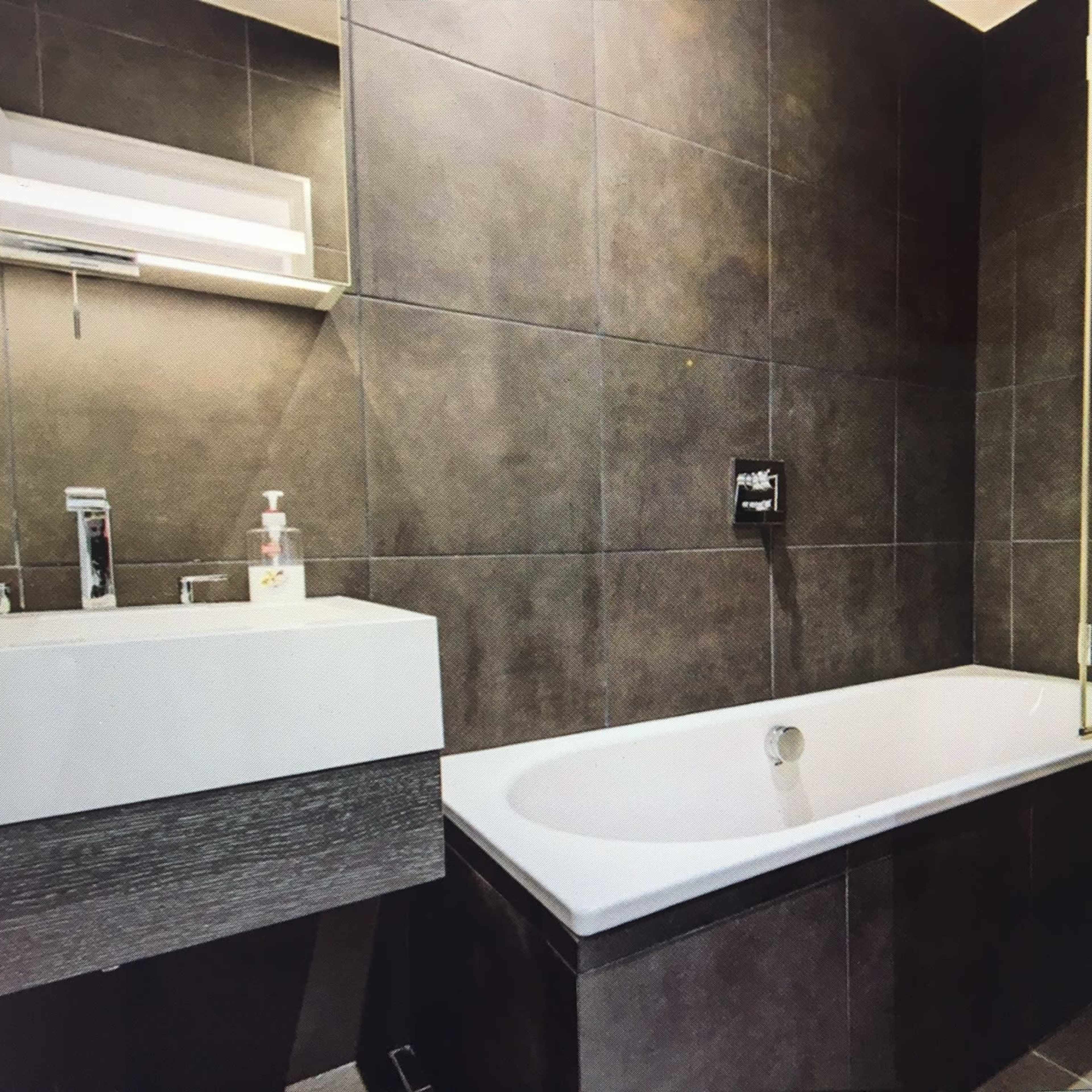 A modern bathroom featuring a white bathtub, a wall-mounted sink with a dark vanity, and a toilet, all set against dark tiled walls.