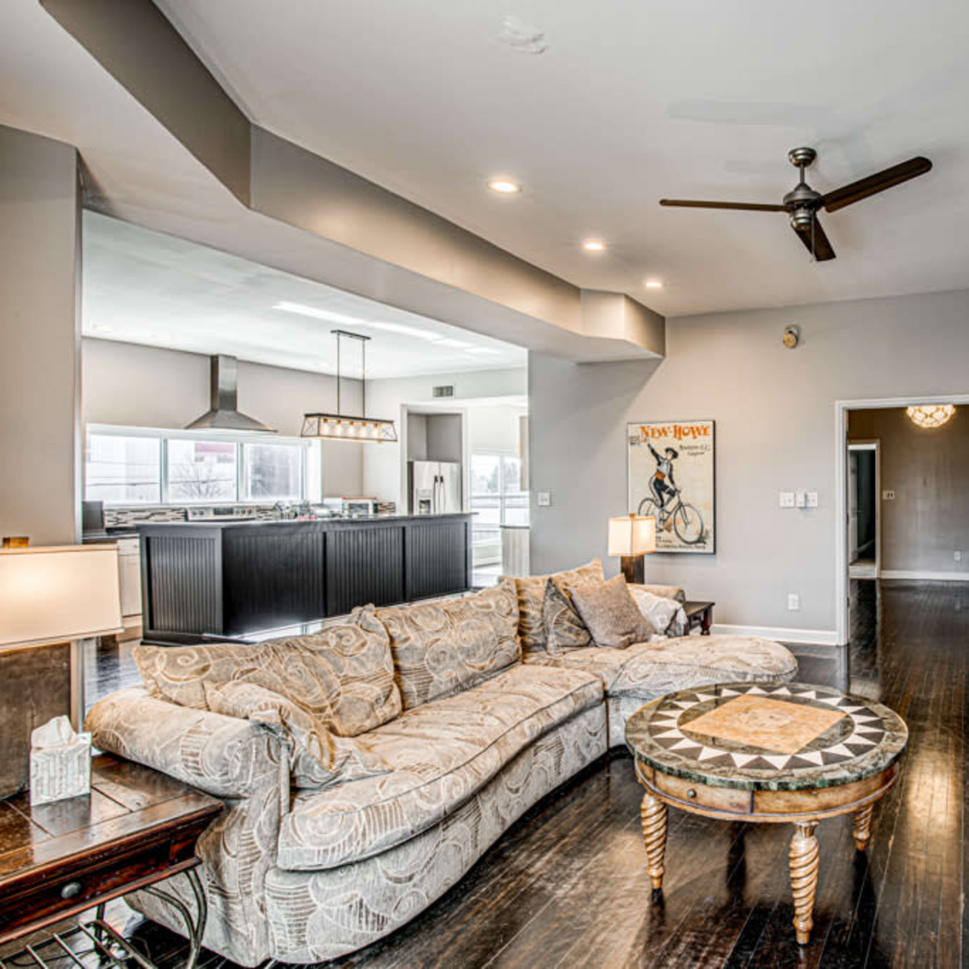 The living room features a large sectional sofa, a decorative coffee table, and an open layout leading to a kitchen area in the background.
