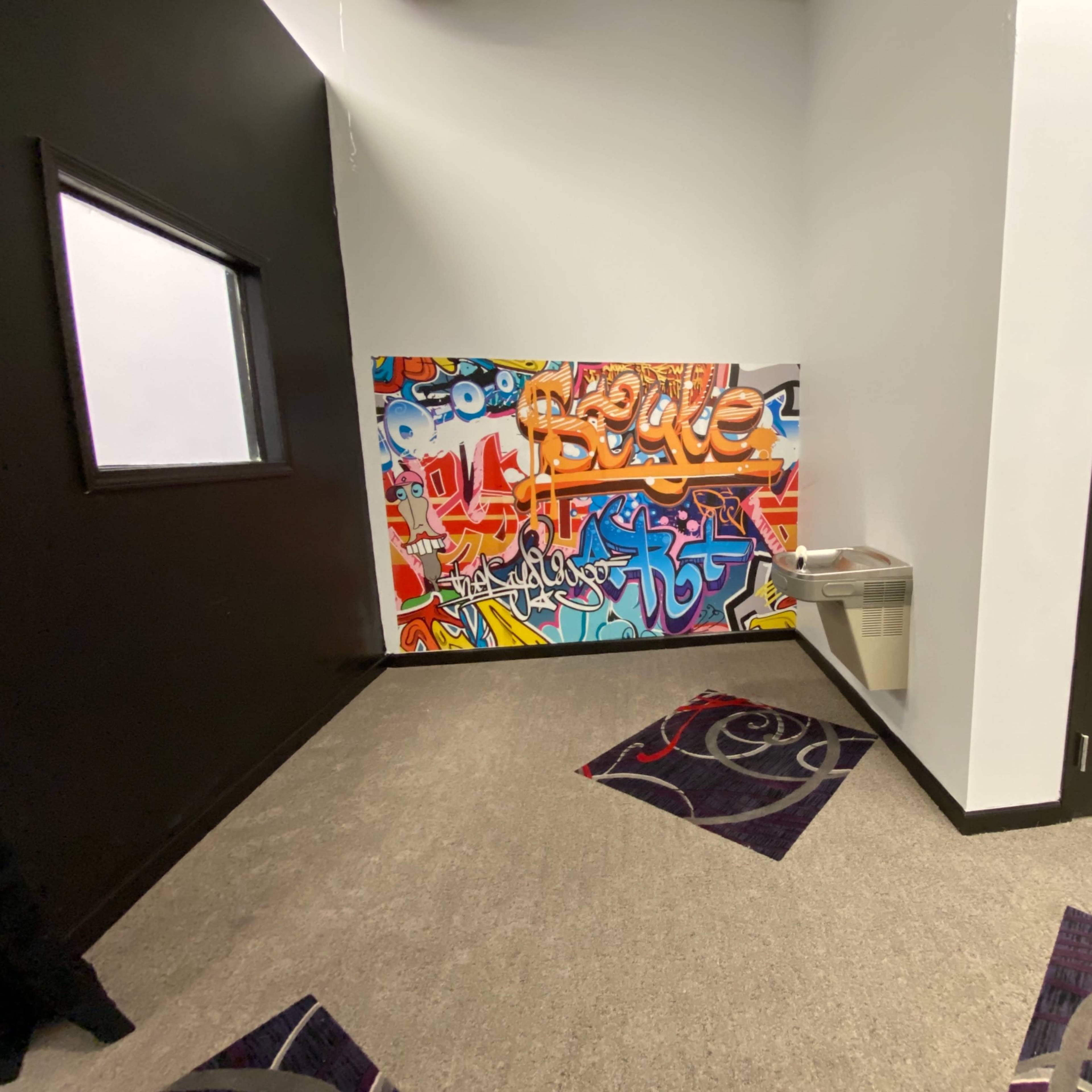 A corridor with a colorful graffiti mural on the wall, a trash can beside it, and a restroom door on the right.