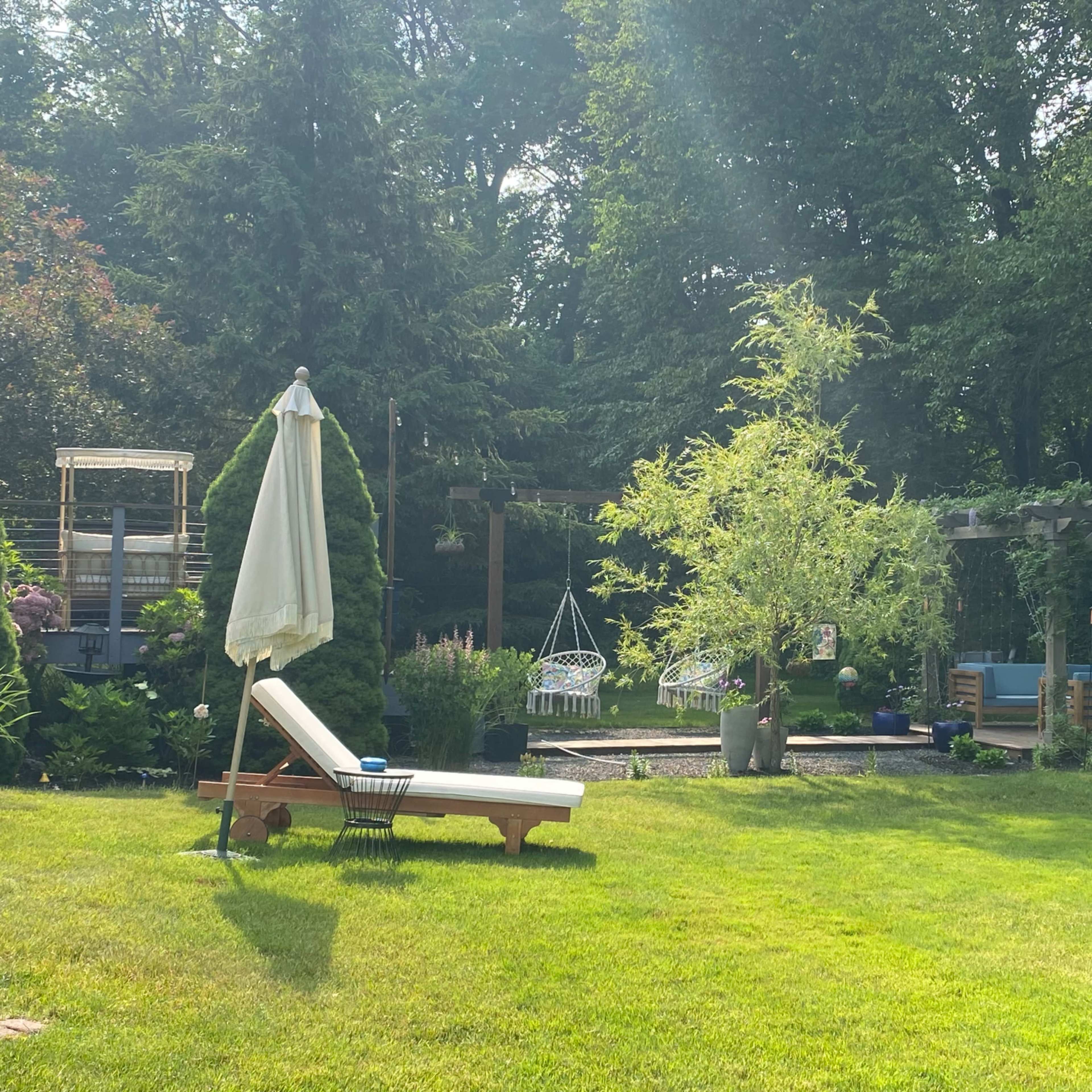 The scene shows a well-kept garden with a lounge chair, an umbrella, swings, and a pergola surrounded by greenery.