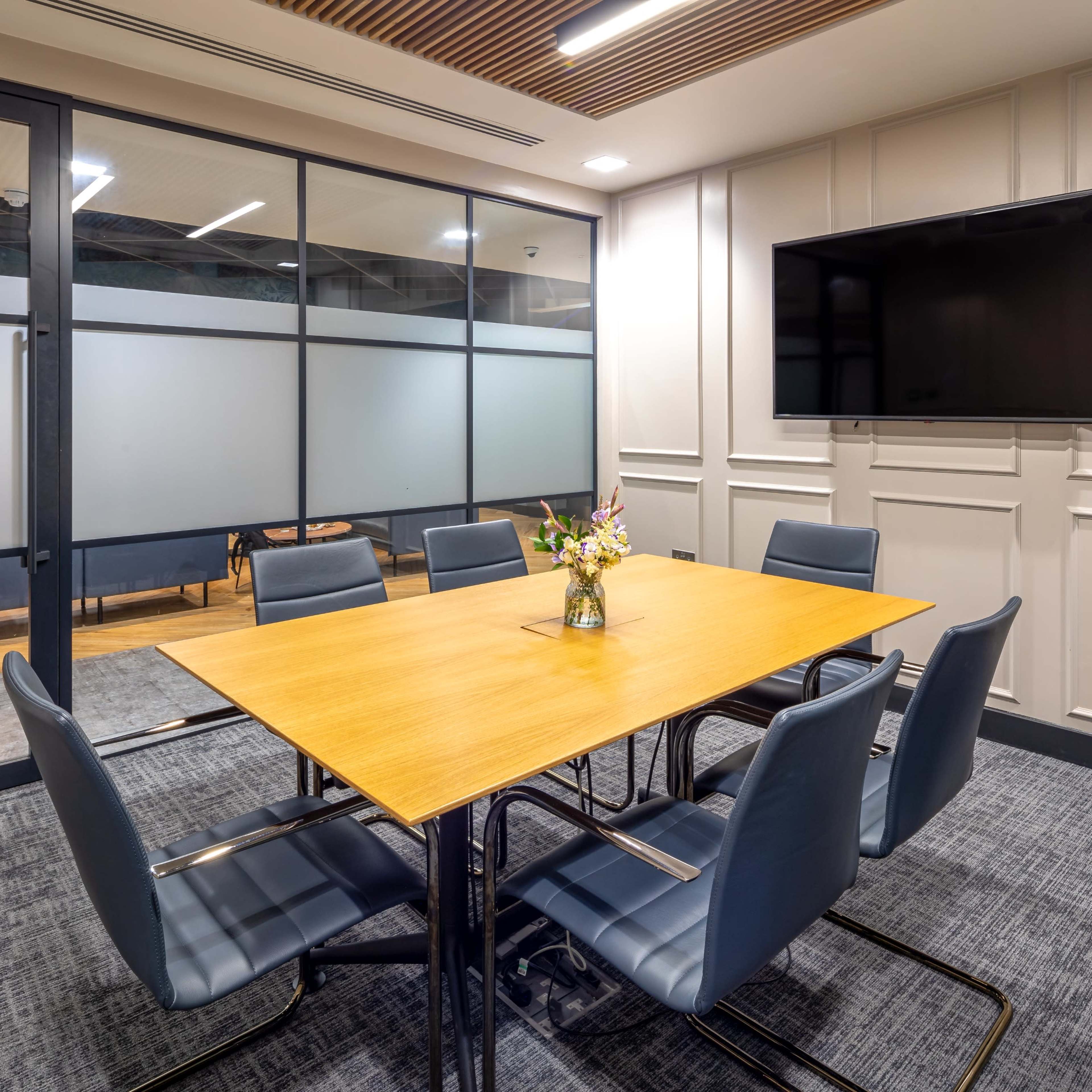 A modern conference room features a large wooden table surrounded by six blue chairs, with a wall-mounted television and a flower vase at the center.
