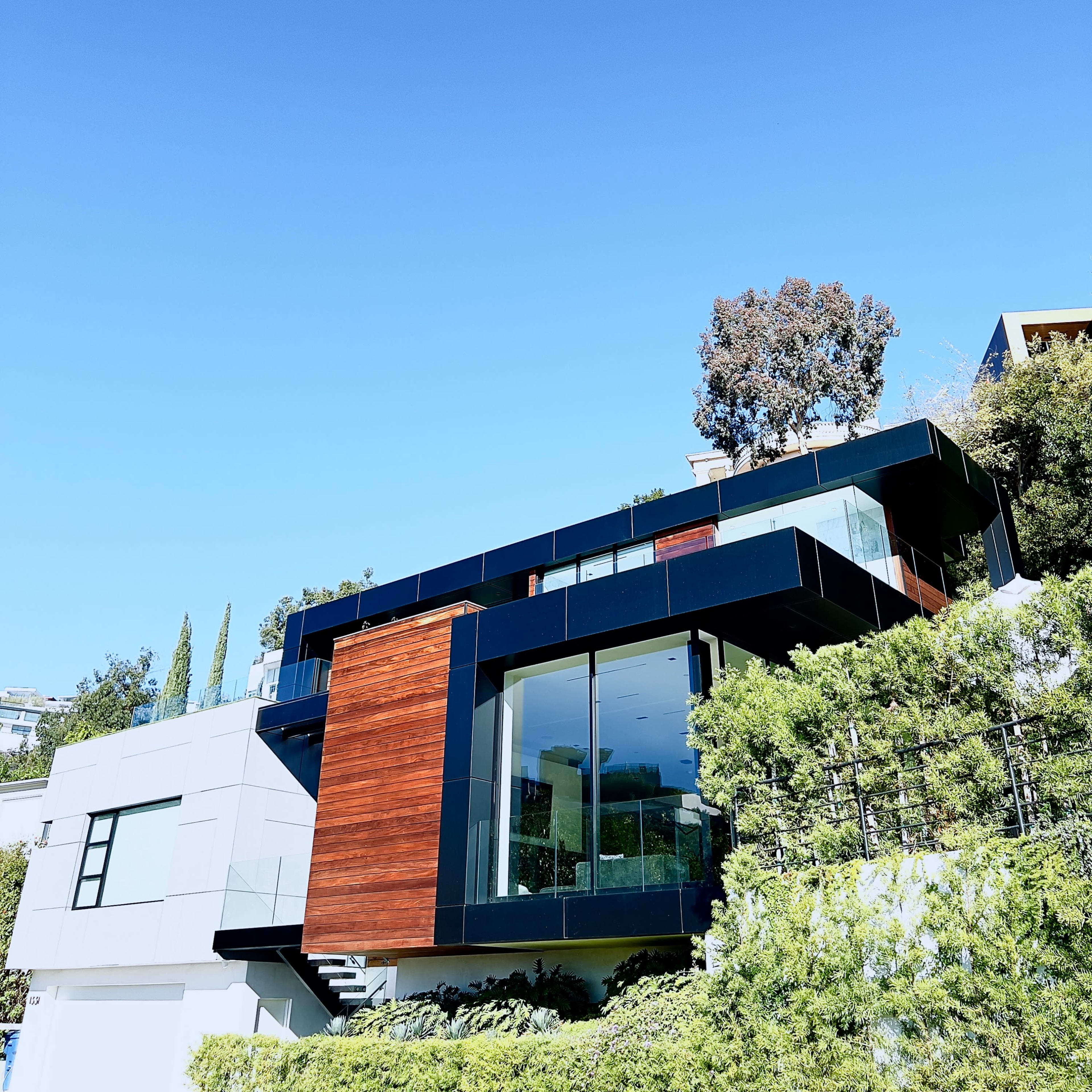 A modern house features large glass windows, wooden accents, and is surrounded by greenery under a clear blue sky.
