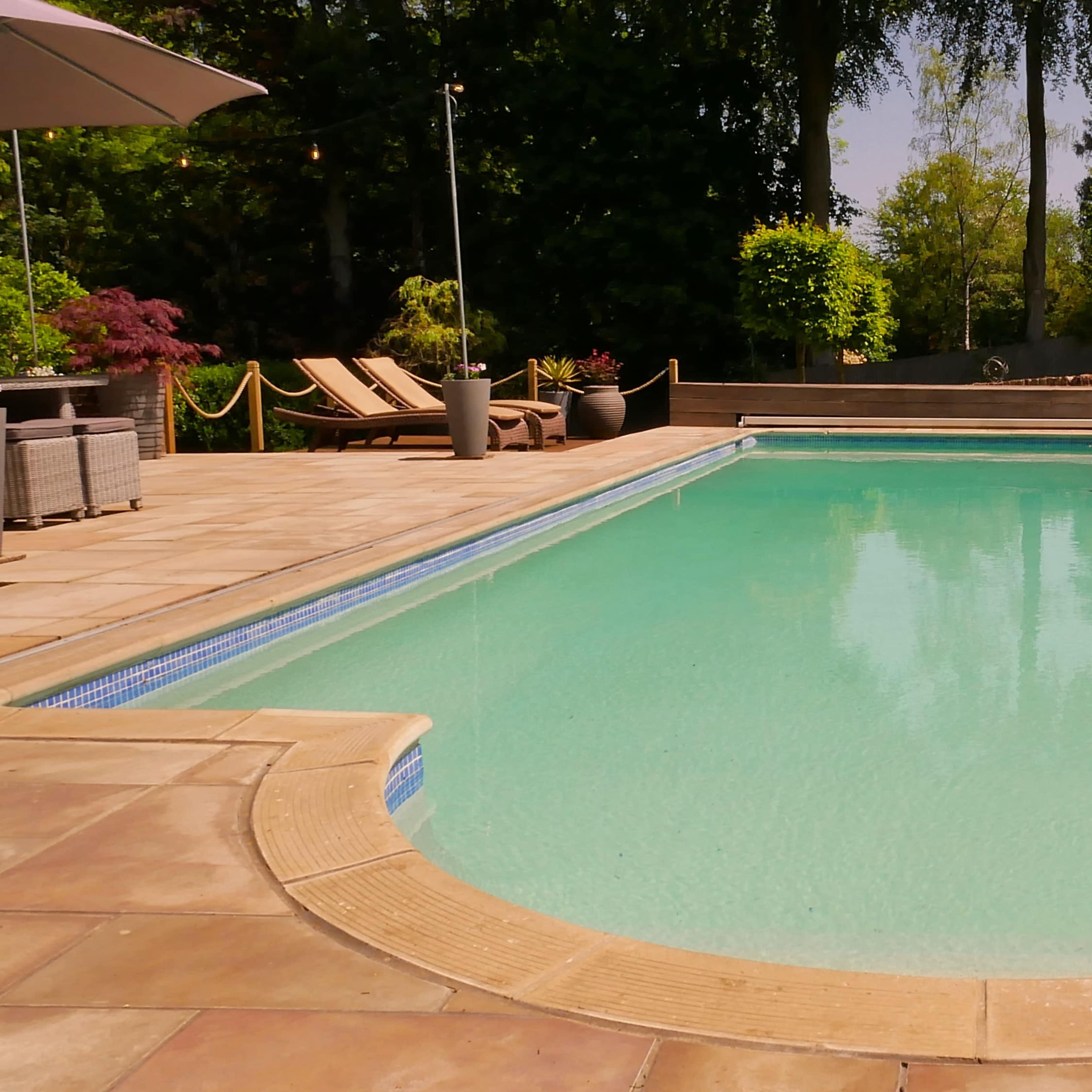 A clear swimming pool is surrounded by a stone patio and landscaped greenery.