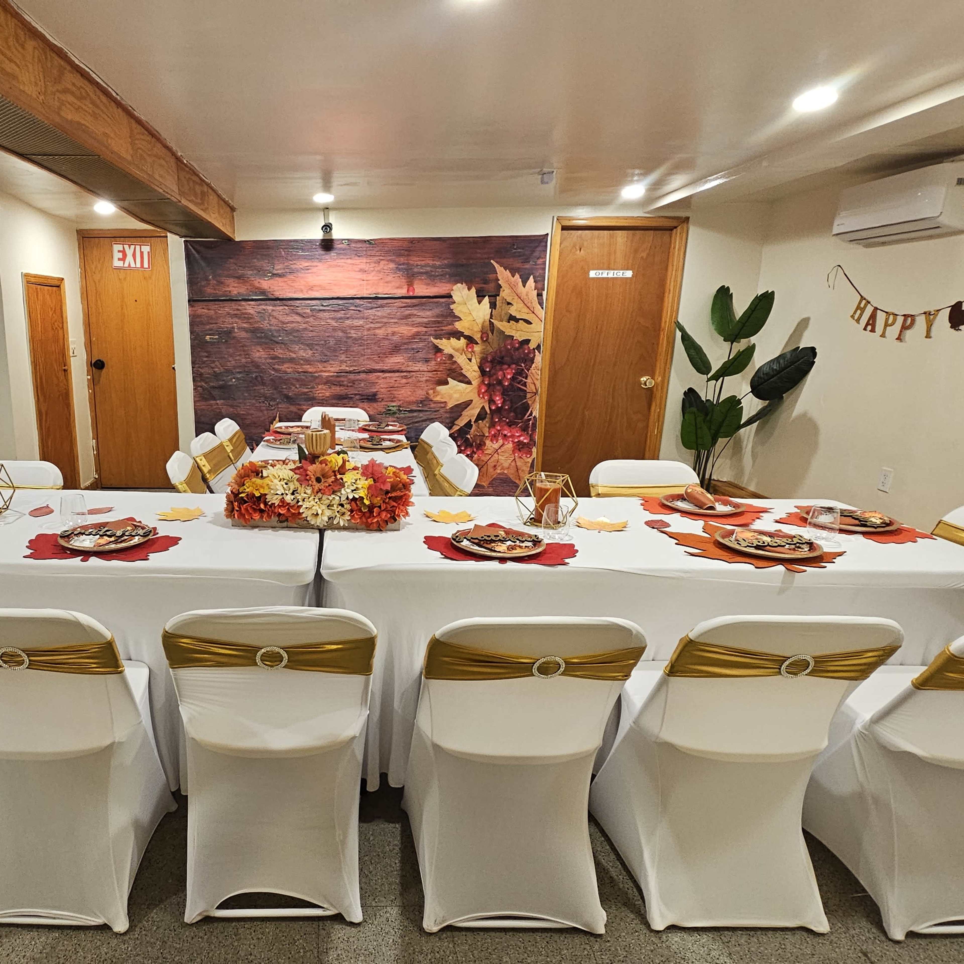 A long table is set for a festive meal in a decorated event space, with autumn-themed centerpieces and chairs arranged around it.