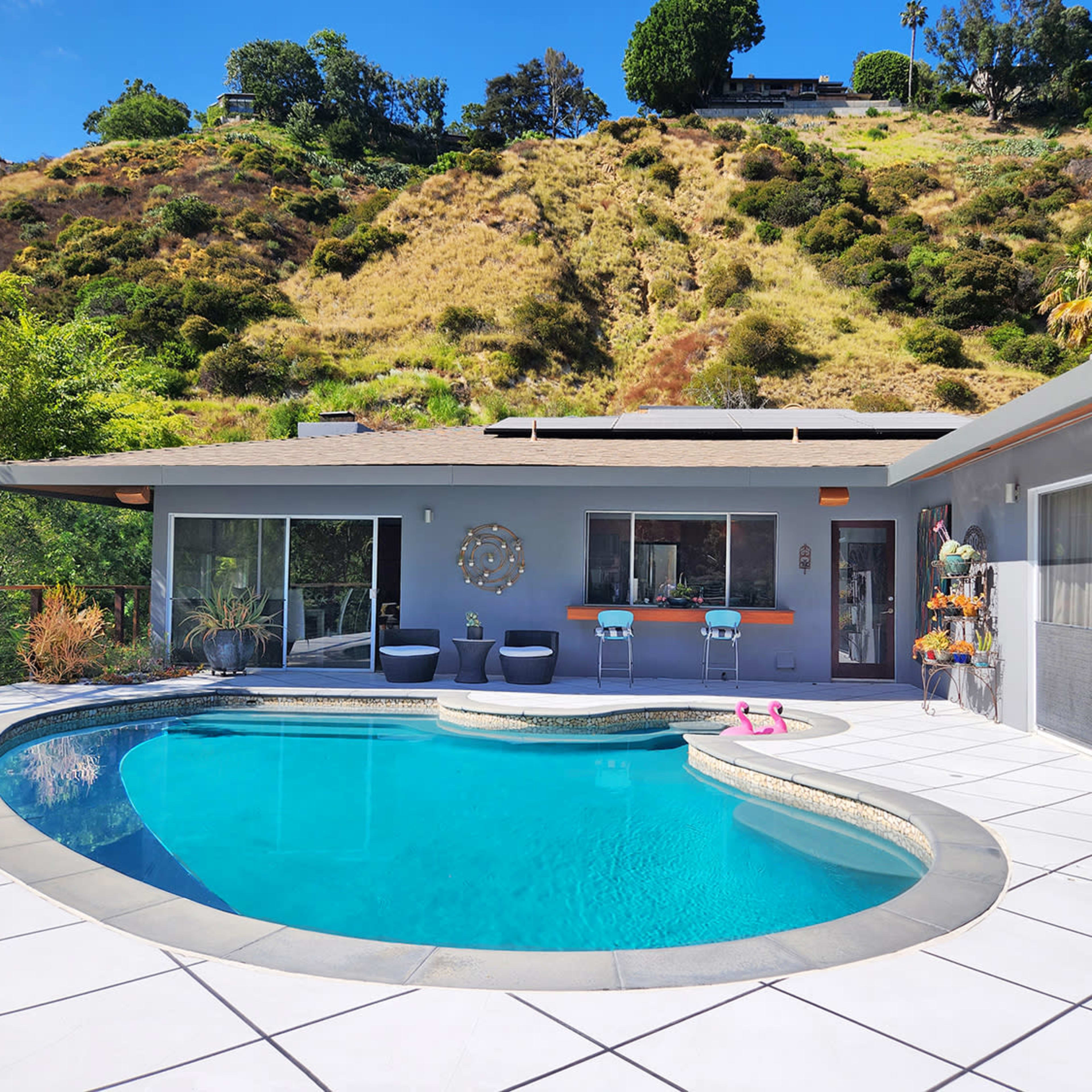 A modern house with a swimming pool, situated on a hillside with green vegetation in the background.