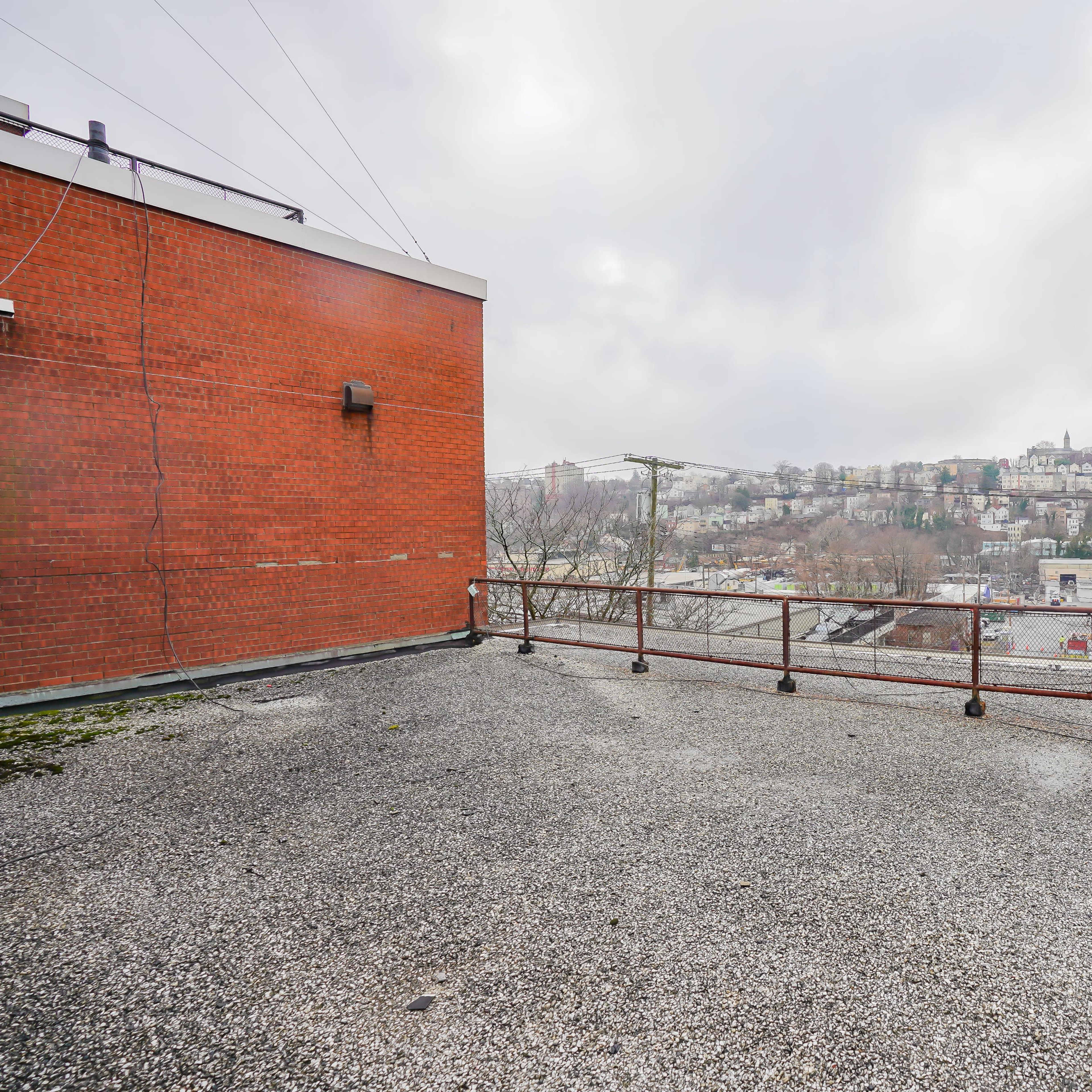 A rooftop area with a gravel surface and a railing overlooking a hillside populated with buildings and trees under a cloudy sky.