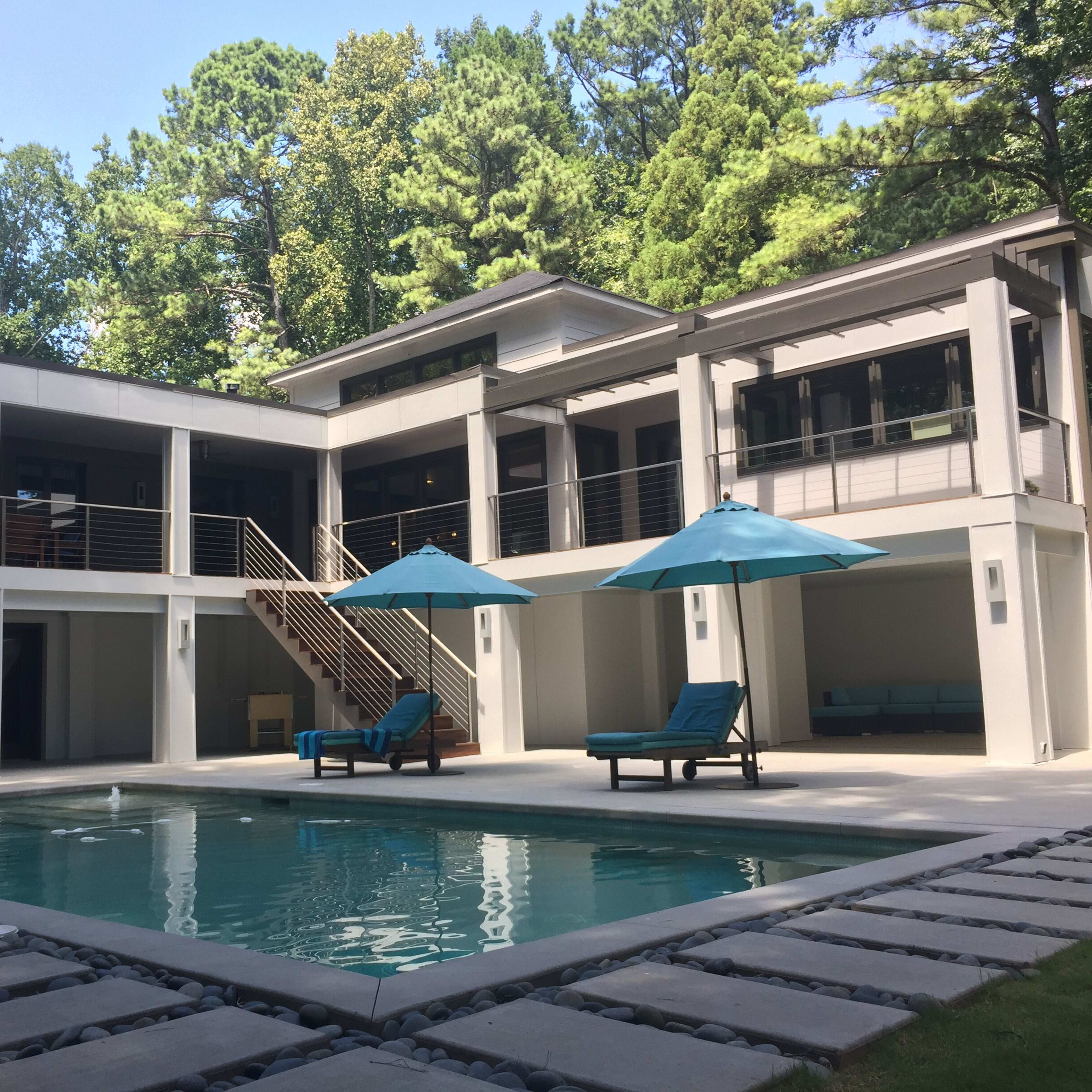 A modern two-story house with a pool, surrounded by trees, featuring outdoor seating areas and umbrellas.