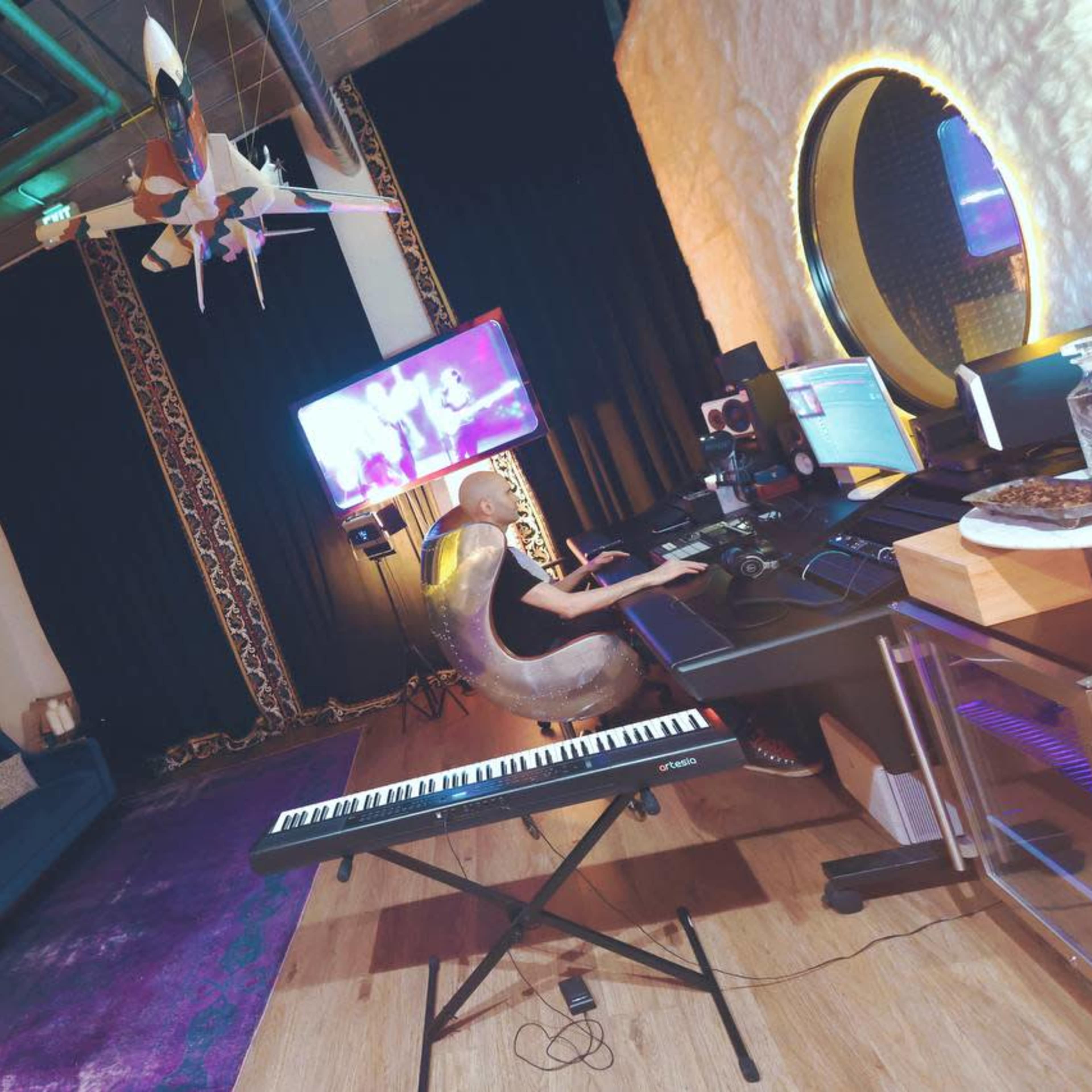 A person is sitting at a workstation with a keyboard and multiple screens in a modern studio featuring a sofa, decorative wall, and large monitor.