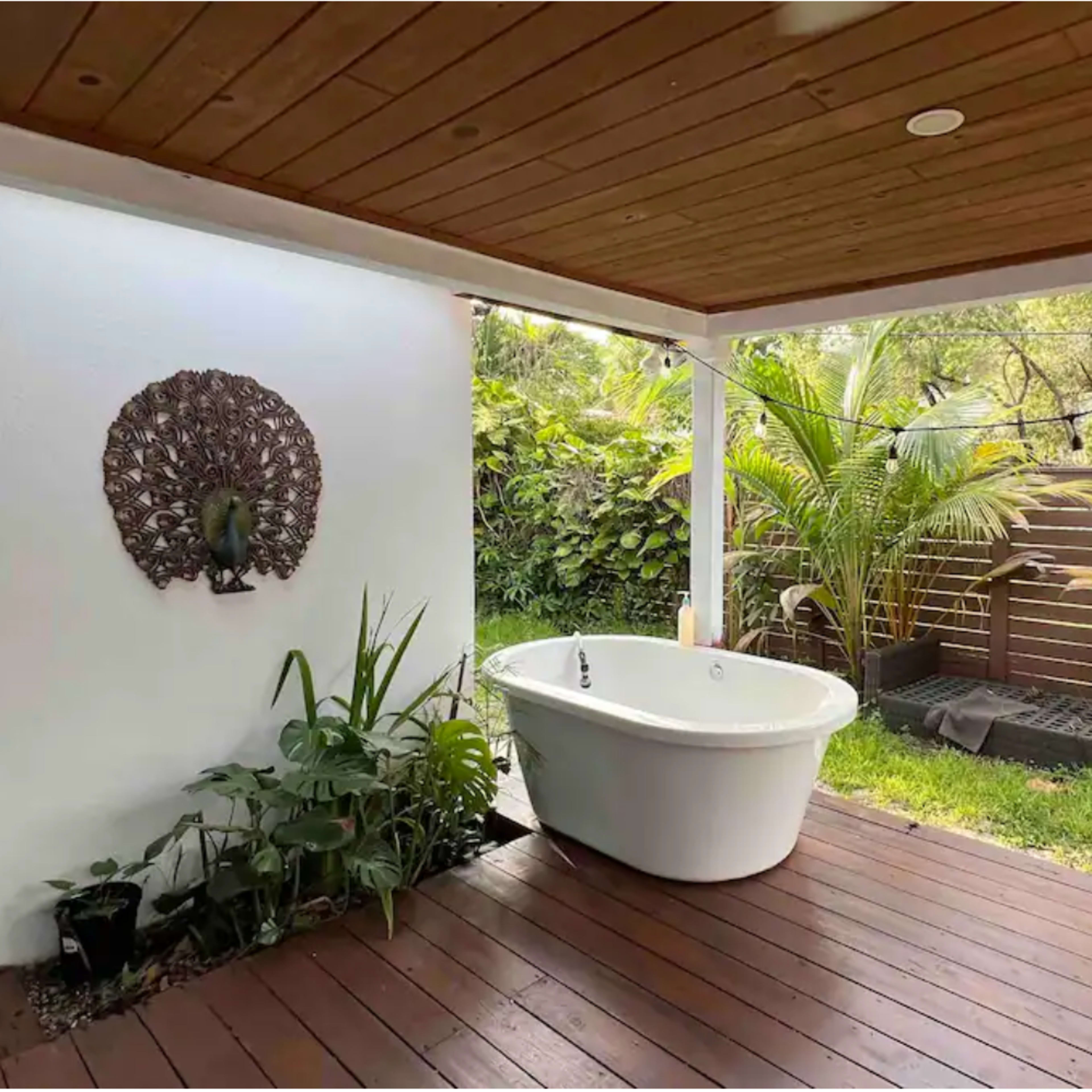 A patio area featuring a freestanding white bathtub surrounded by plants and a wooden deck.