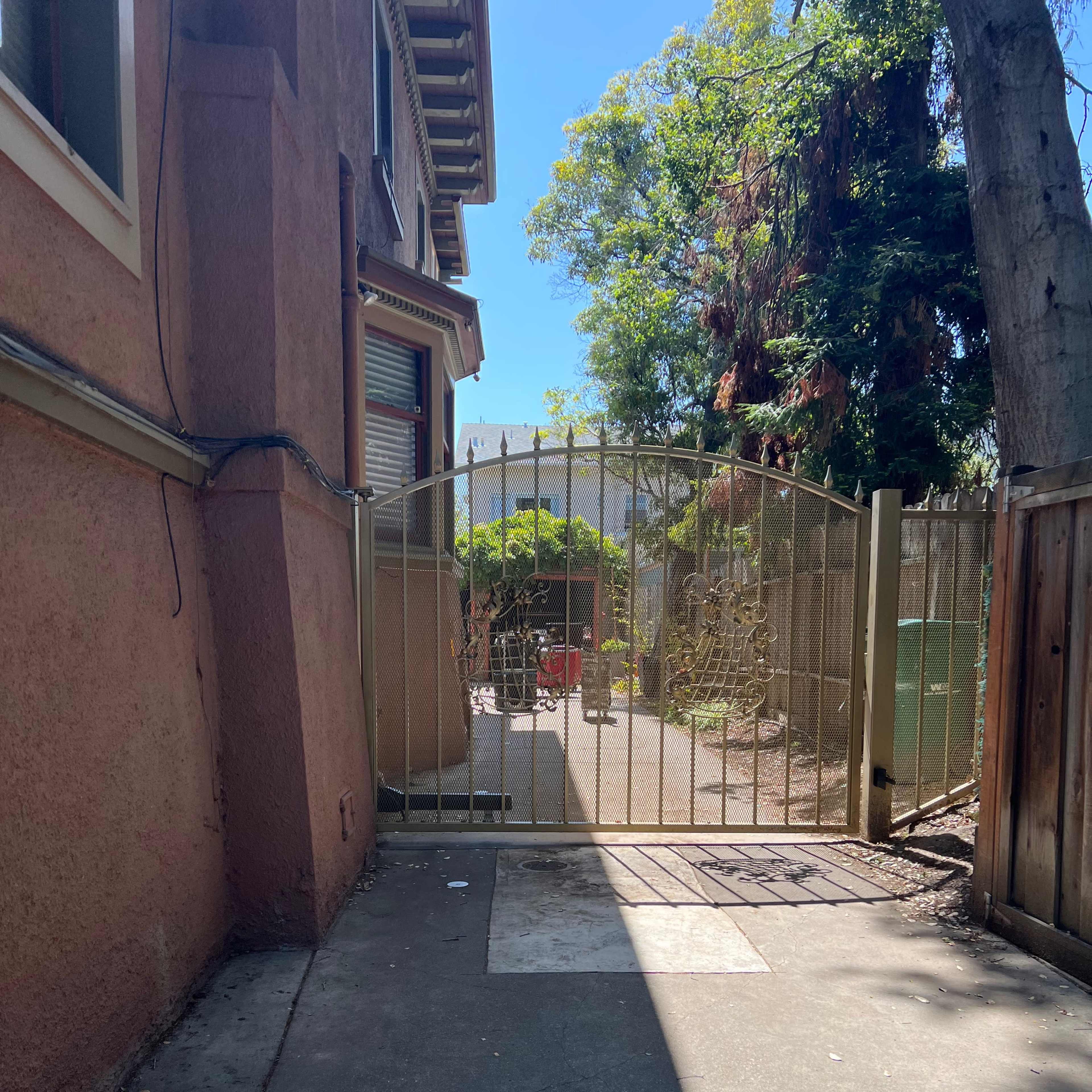 A gated entrance leads to a pathway beside a building, surrounded by trees and a fence.