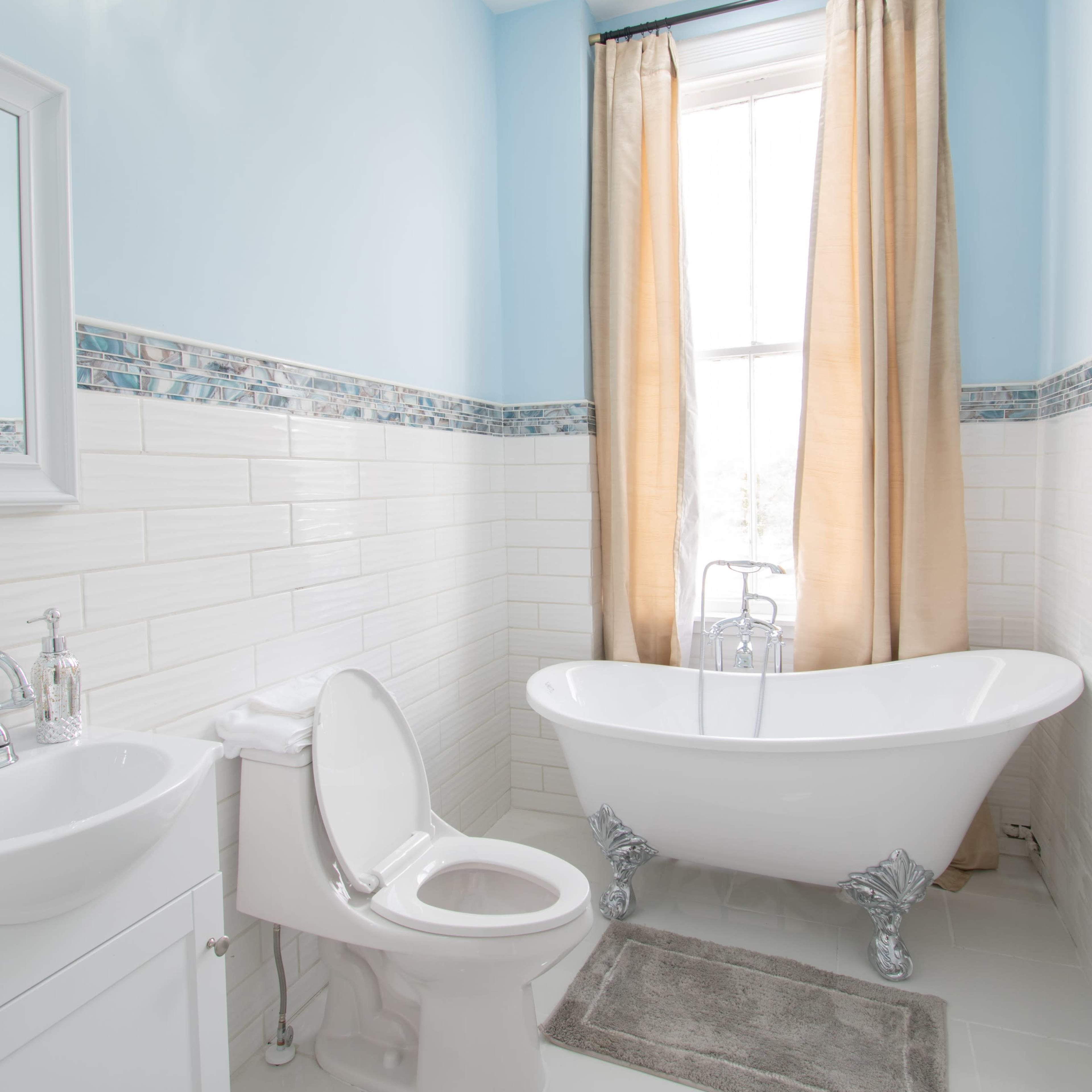 A modern bathroom features a freestanding bathtub, a toilet, a sink with a mirror, and large windows with beige curtains.