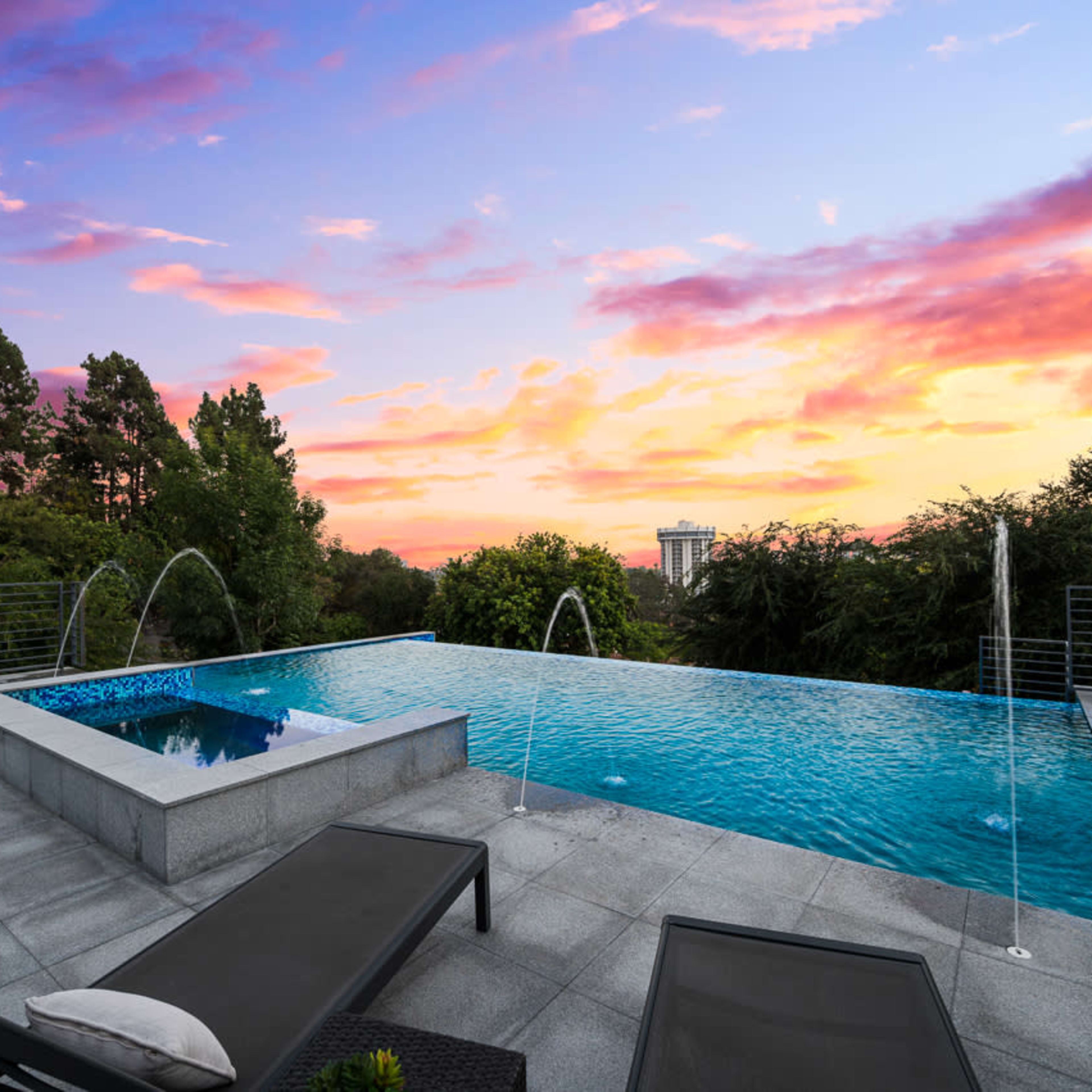 A modern swimming pool with an infinity edge, surrounded by trees and a sunset sky.