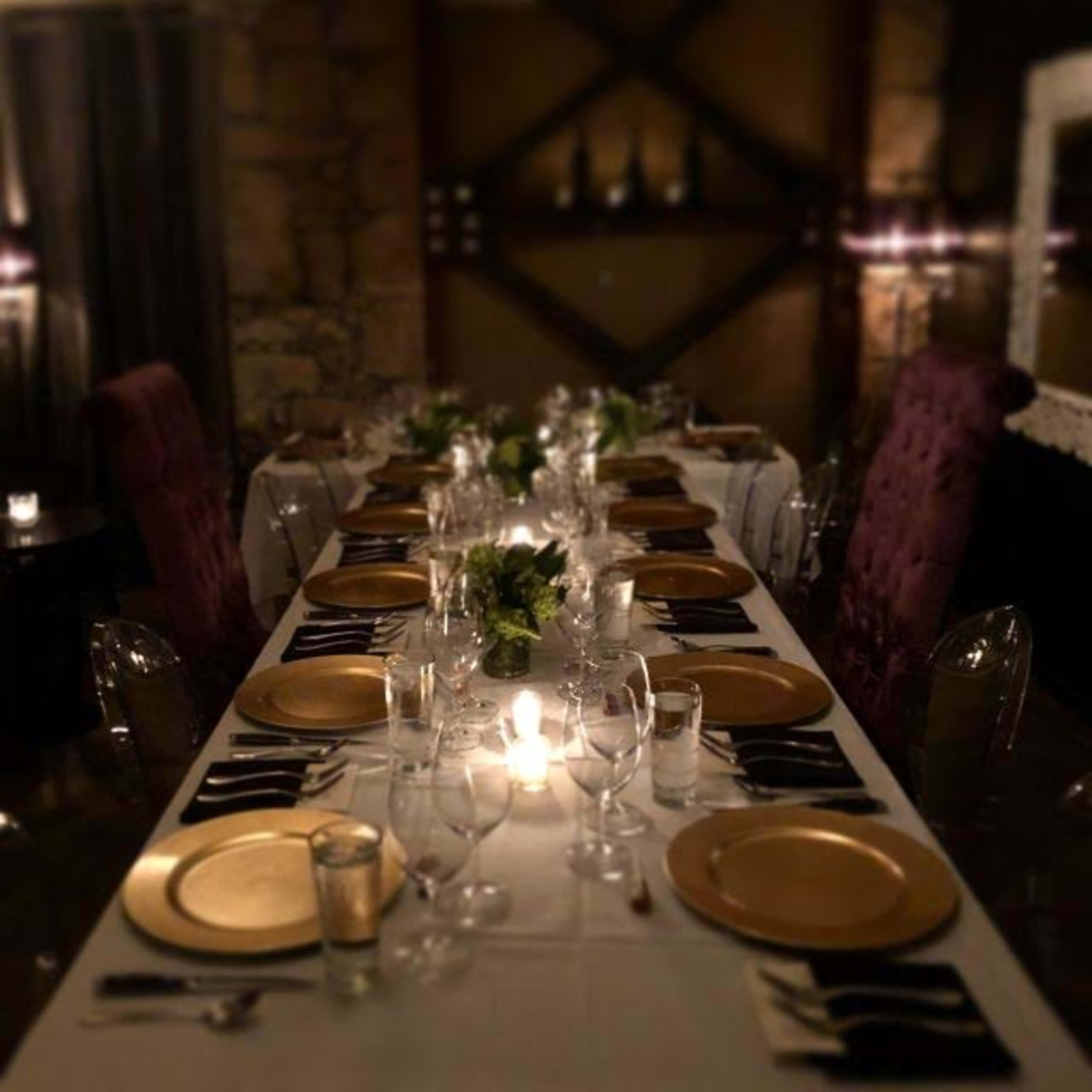 A long dining table is set with gold plates, glassware, and foliage in a dimly lit room.