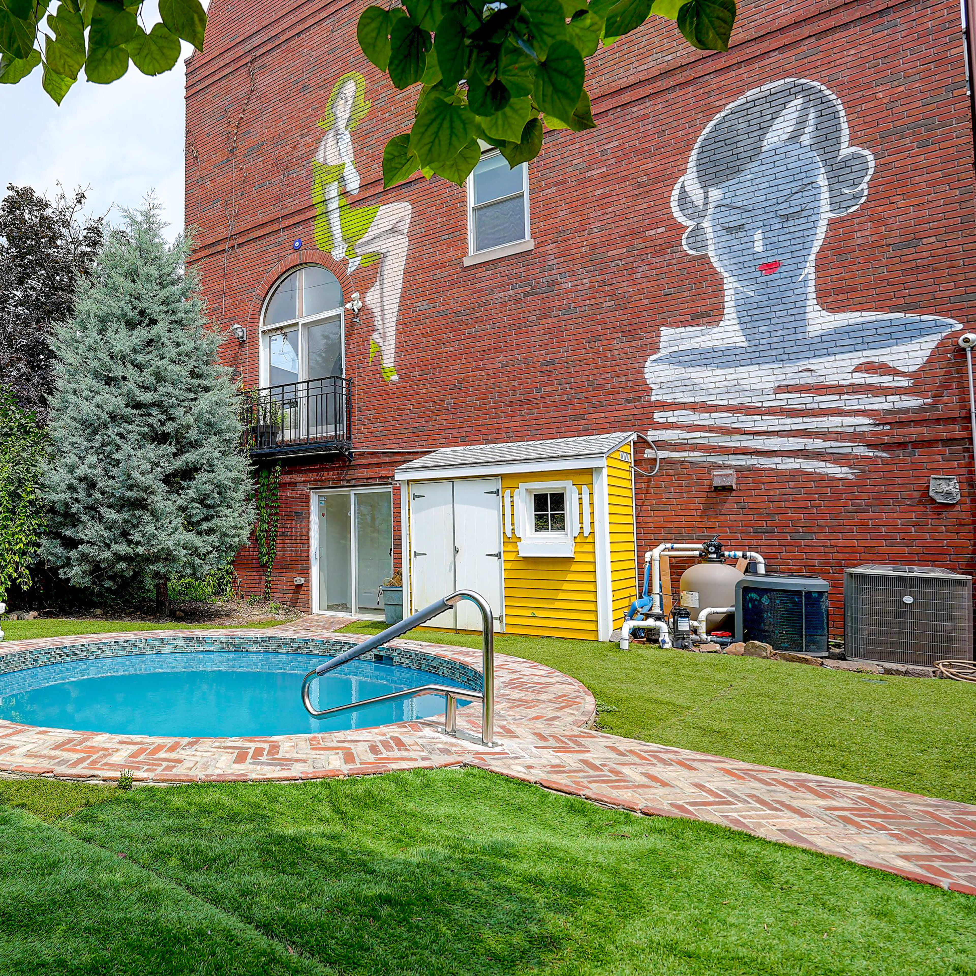 The scene features a small circular pool surrounded by a brick patio and a lush green lawn, with a mural of a woman on the brick wall of a building in the background.