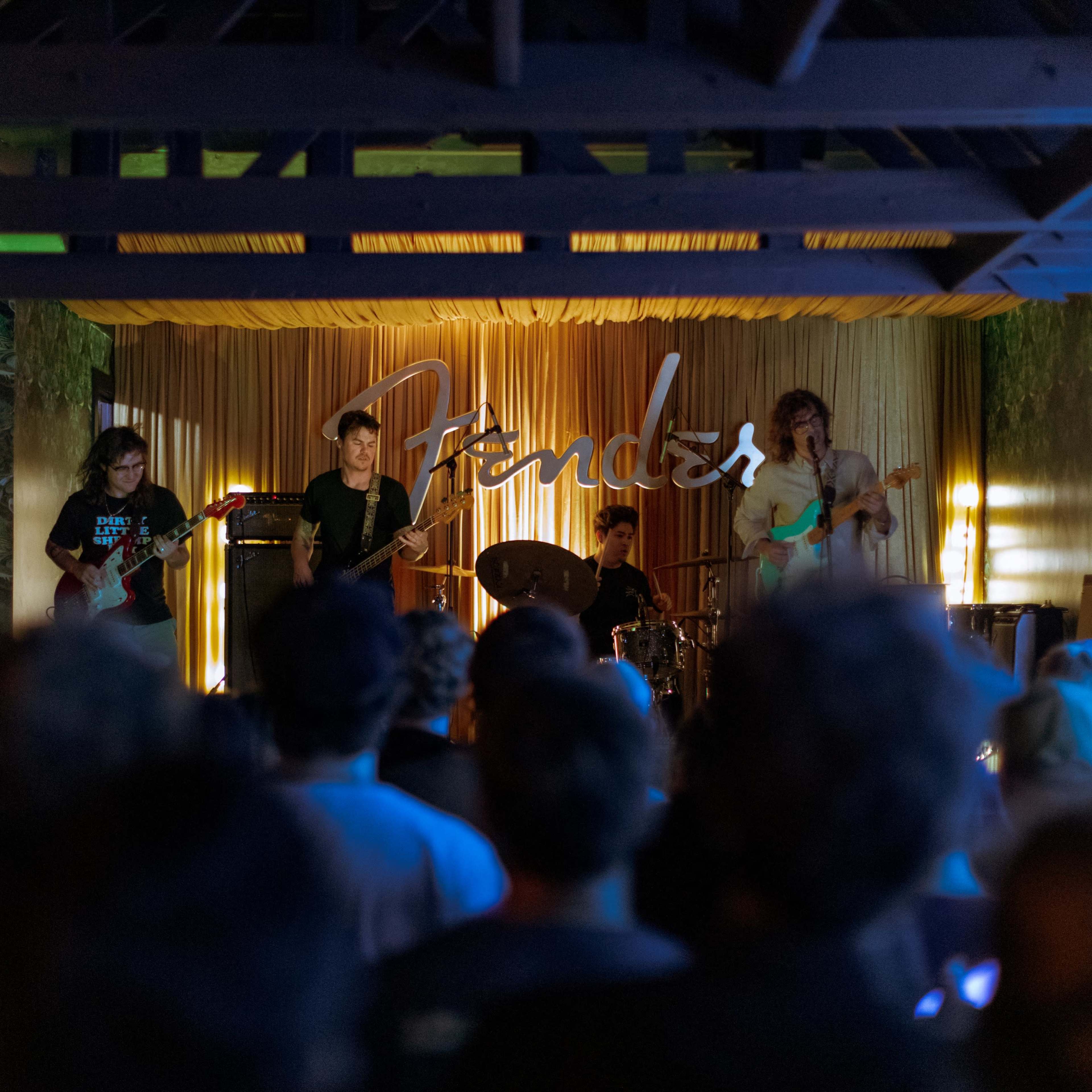 A band performs on stage in front of an audience, with a large Fender logo displayed prominently behind them.