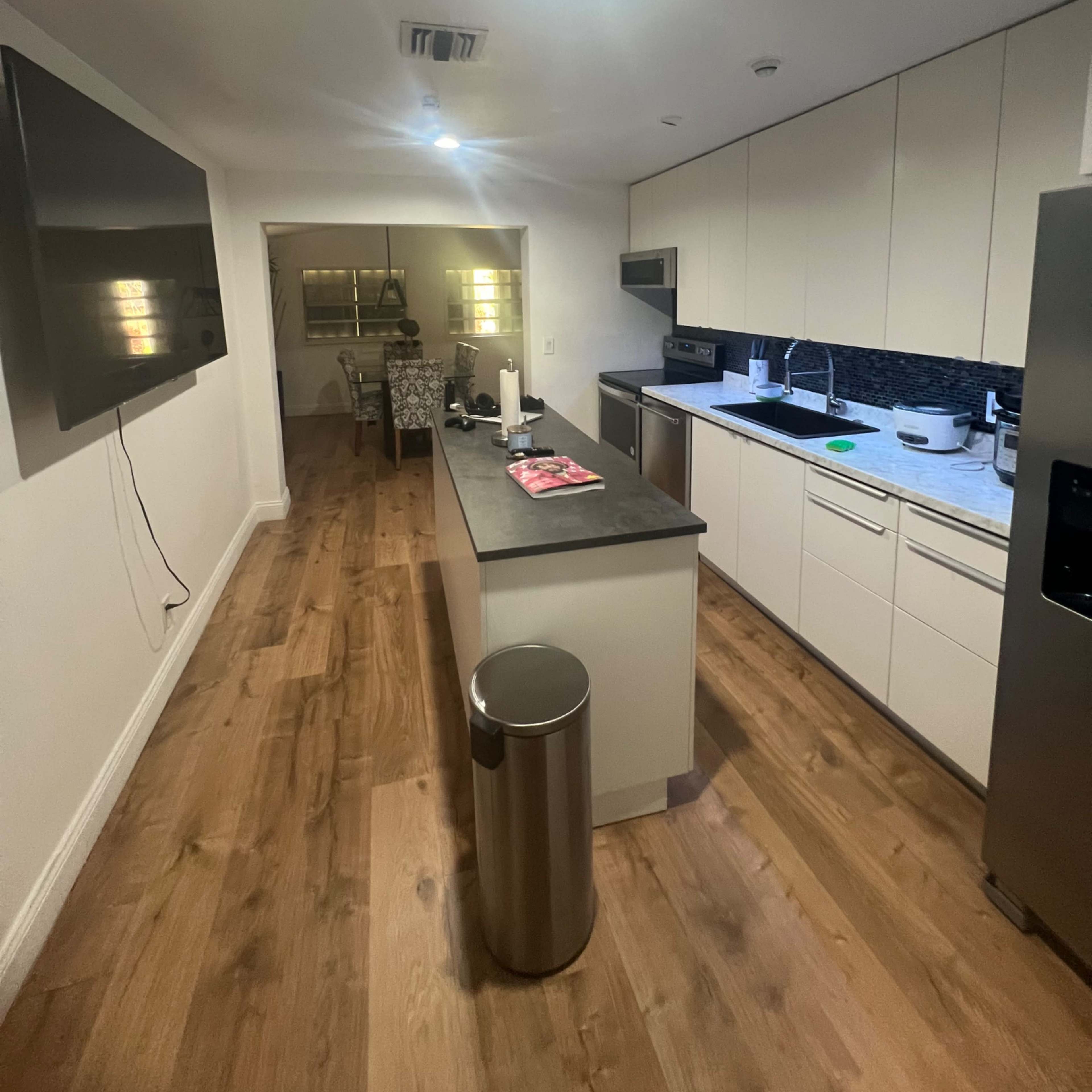 A modern kitchen with a central island, equipped with appliances, a television mounted on the wall, and wooden flooring.