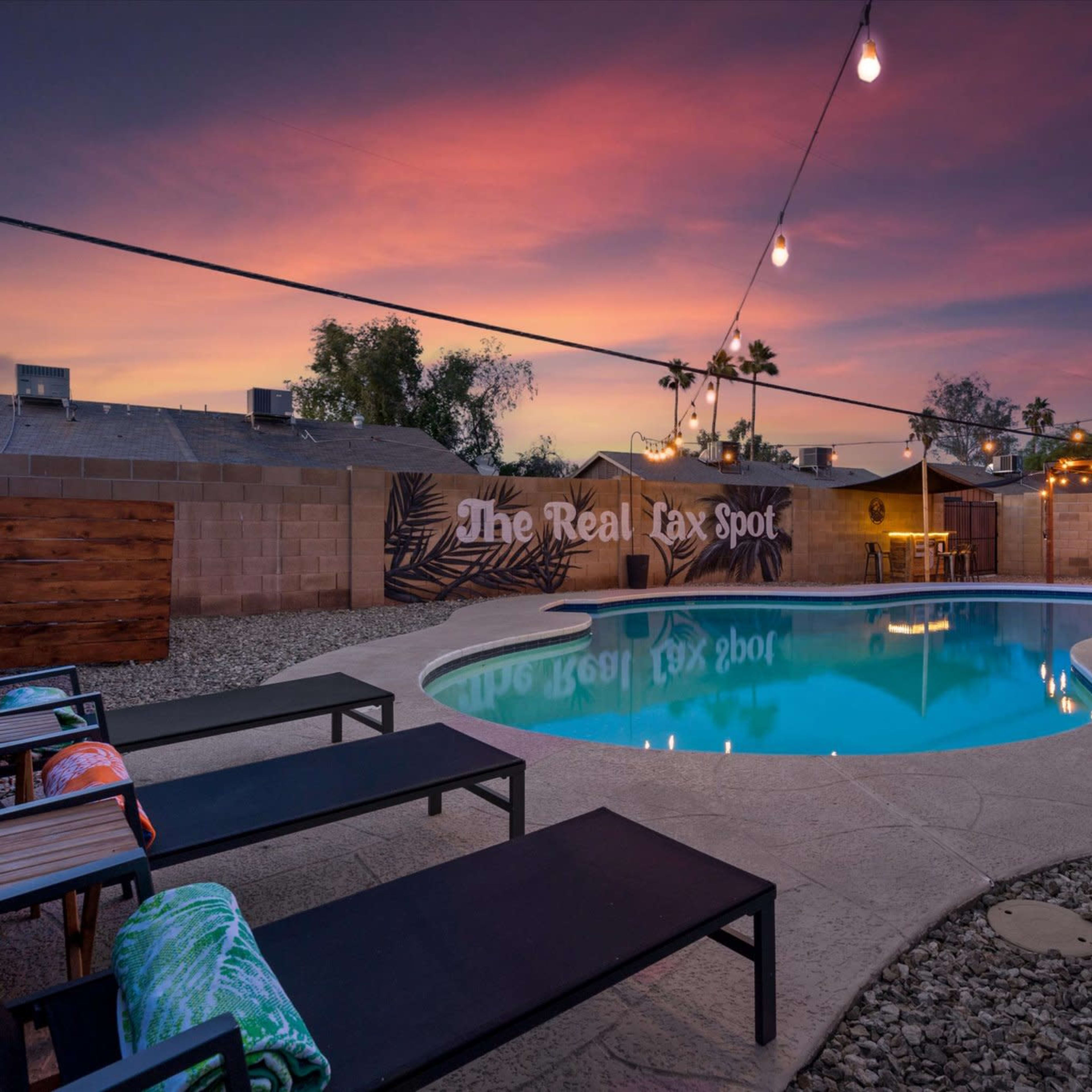 A swimming pool surrounded by lounge chairs and outdoor lighting, set against a colorful sunset sky.