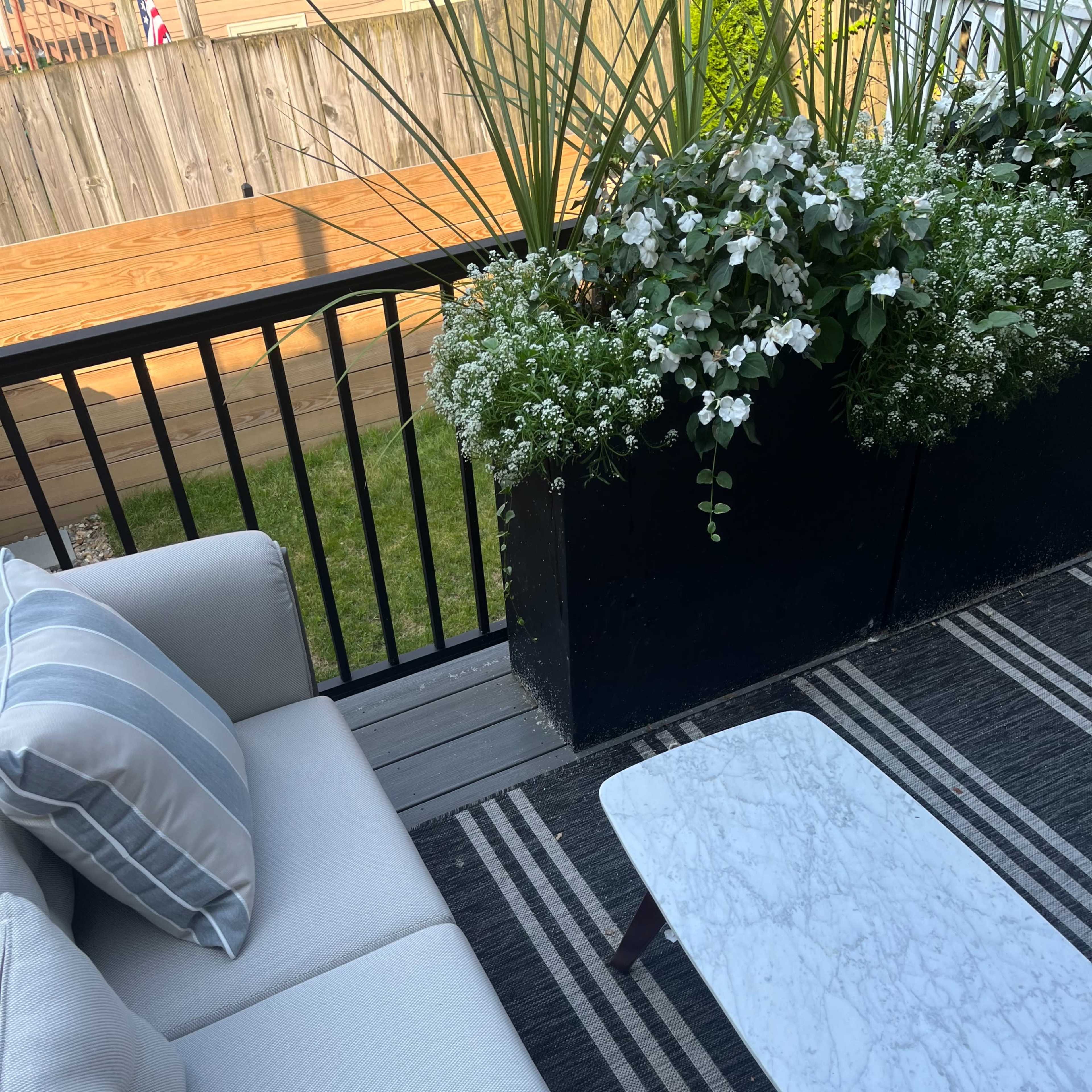 A patio area features a light-colored sectional sofa with decorative pillows, a marble-topped coffee table, and a planter filled with vibrant plants and flowers.