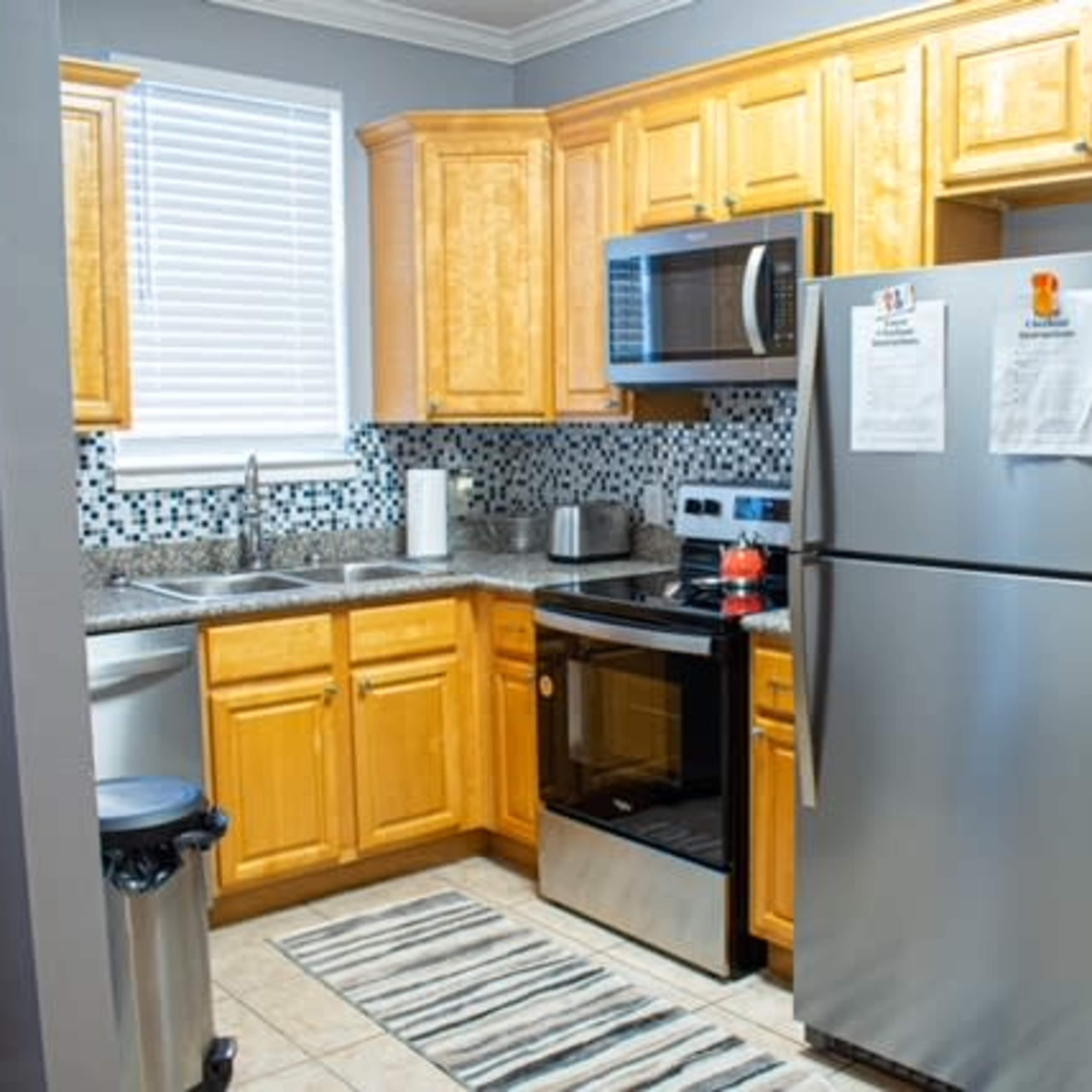 A compact kitchen features wooden cabinets, stainless steel appliances, and a patterned rug on the tiled floor.