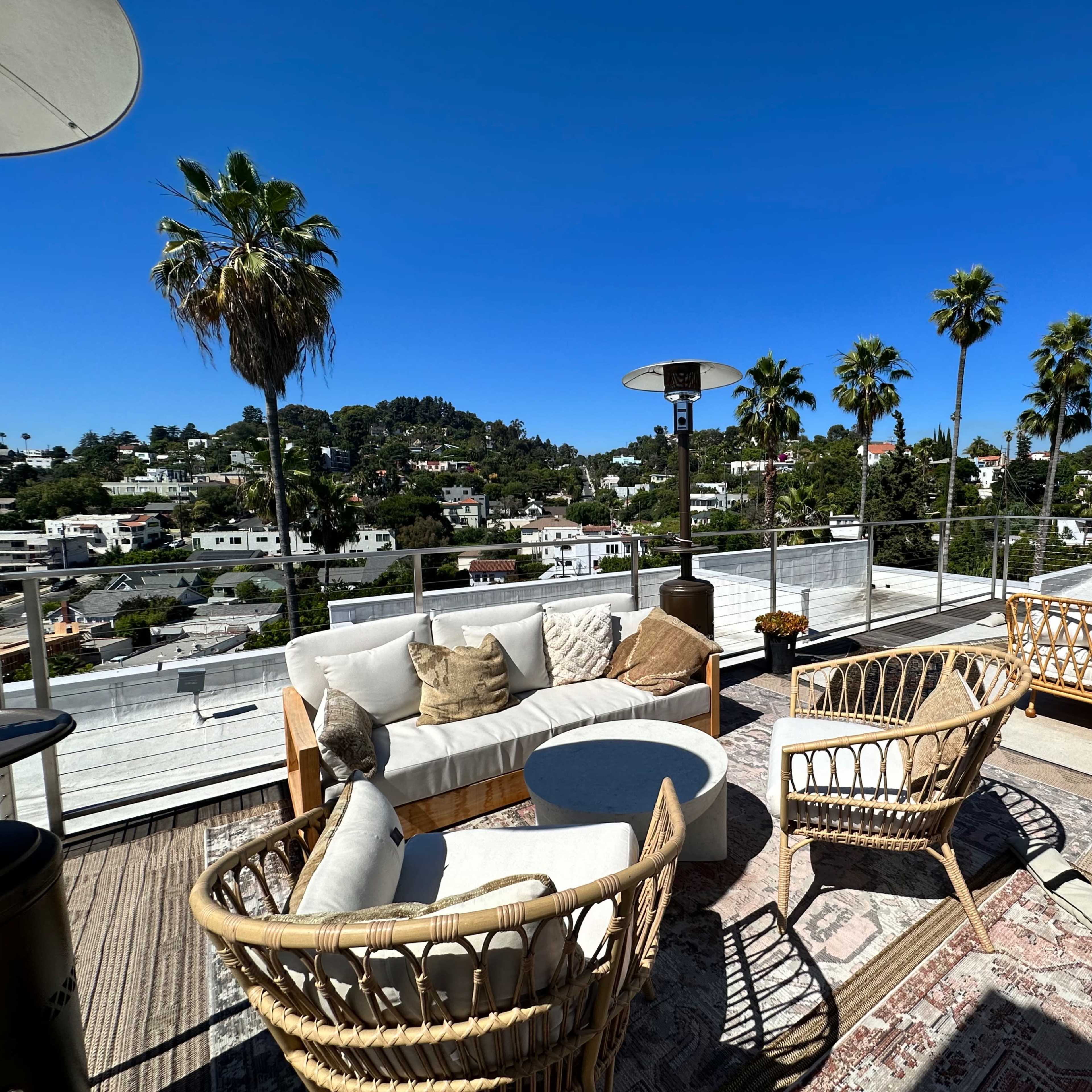 A rooftop terrace features woven furniture, a round table, and palm trees in the background under a clear blue sky.