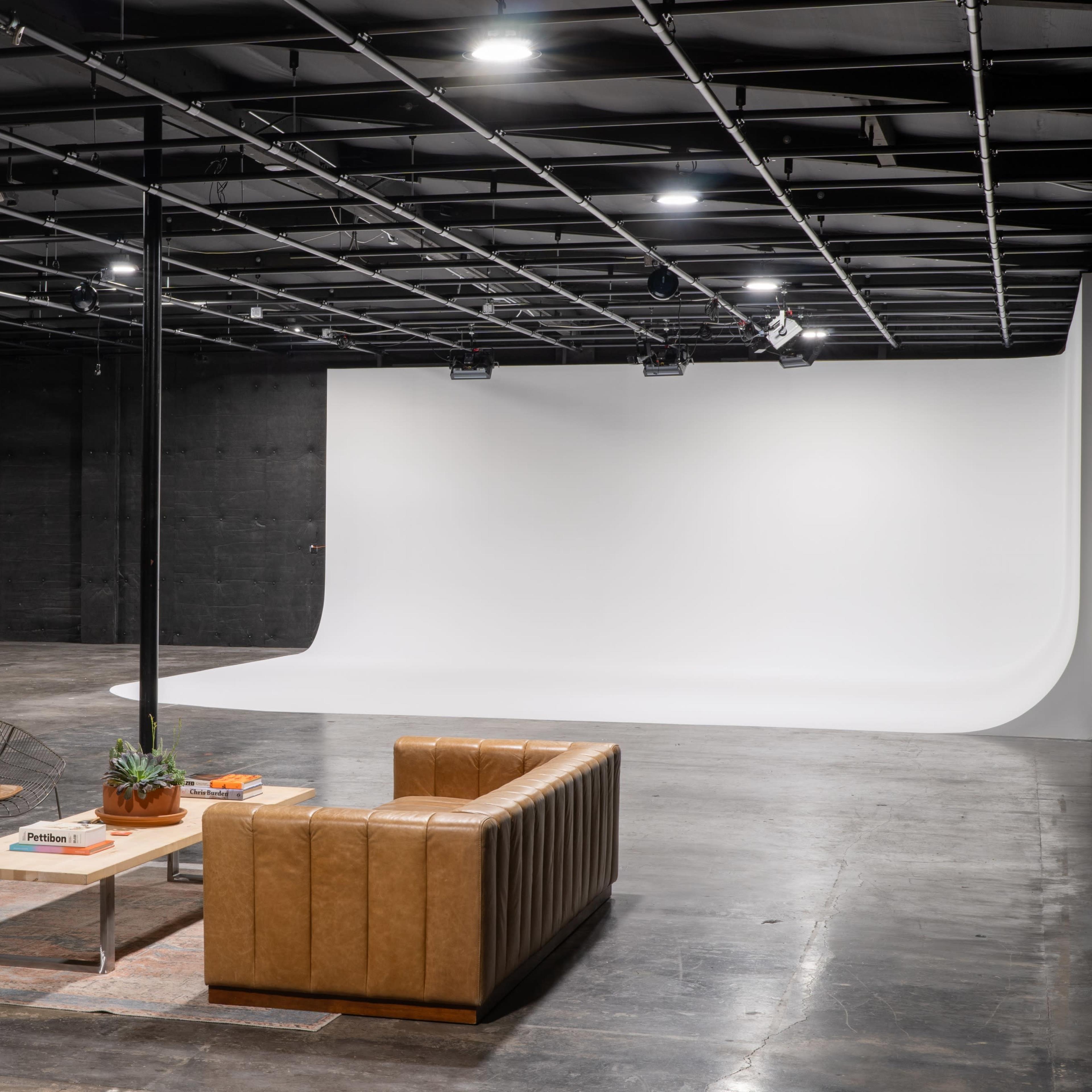 A spacious studio with a minimalist wooden couch, a coffee table, a green chair, and a white backdrop for photography.