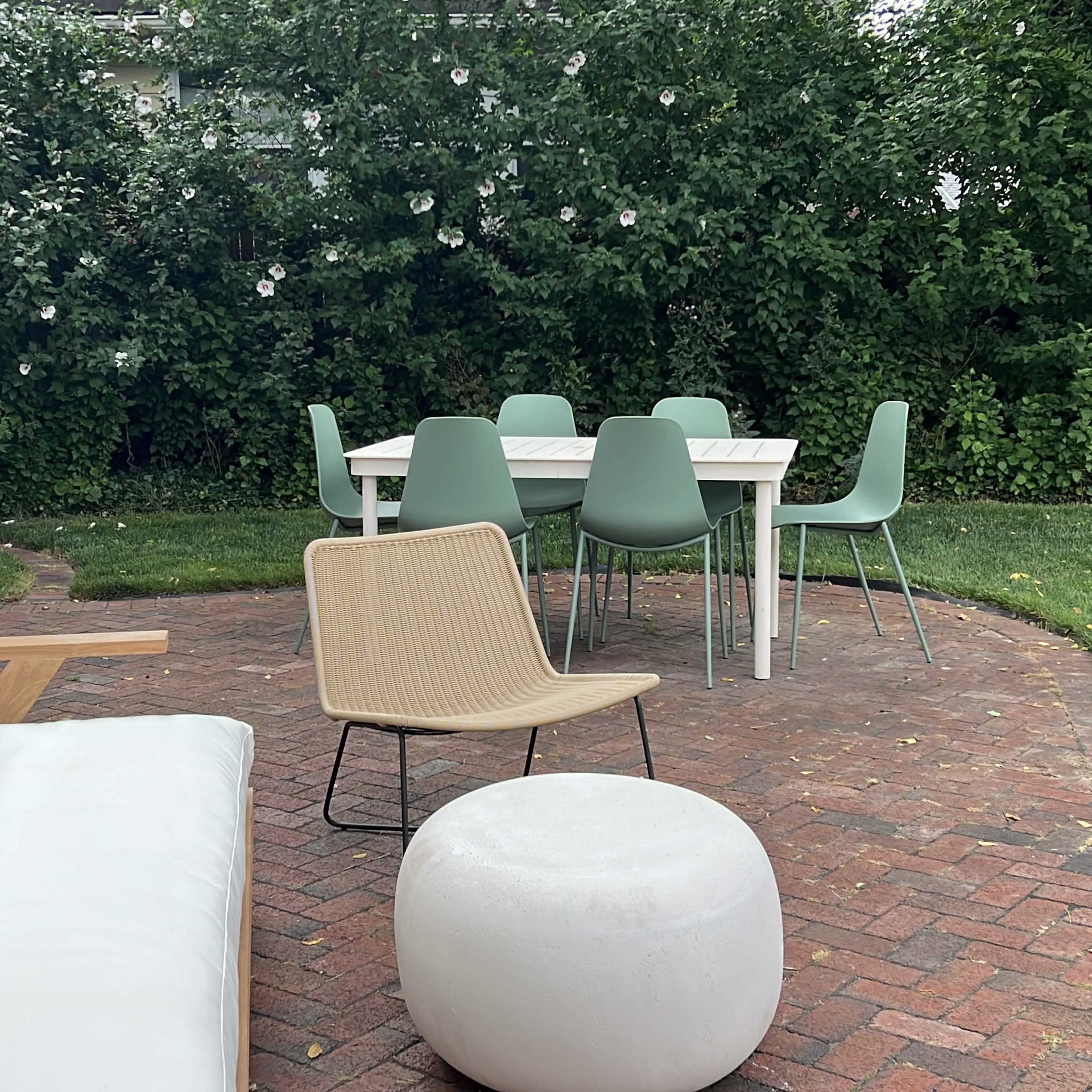 A patio area features a white sofa, a round white pouf, and a dining table surrounded by green chairs, with a backdrop of lush greenery.