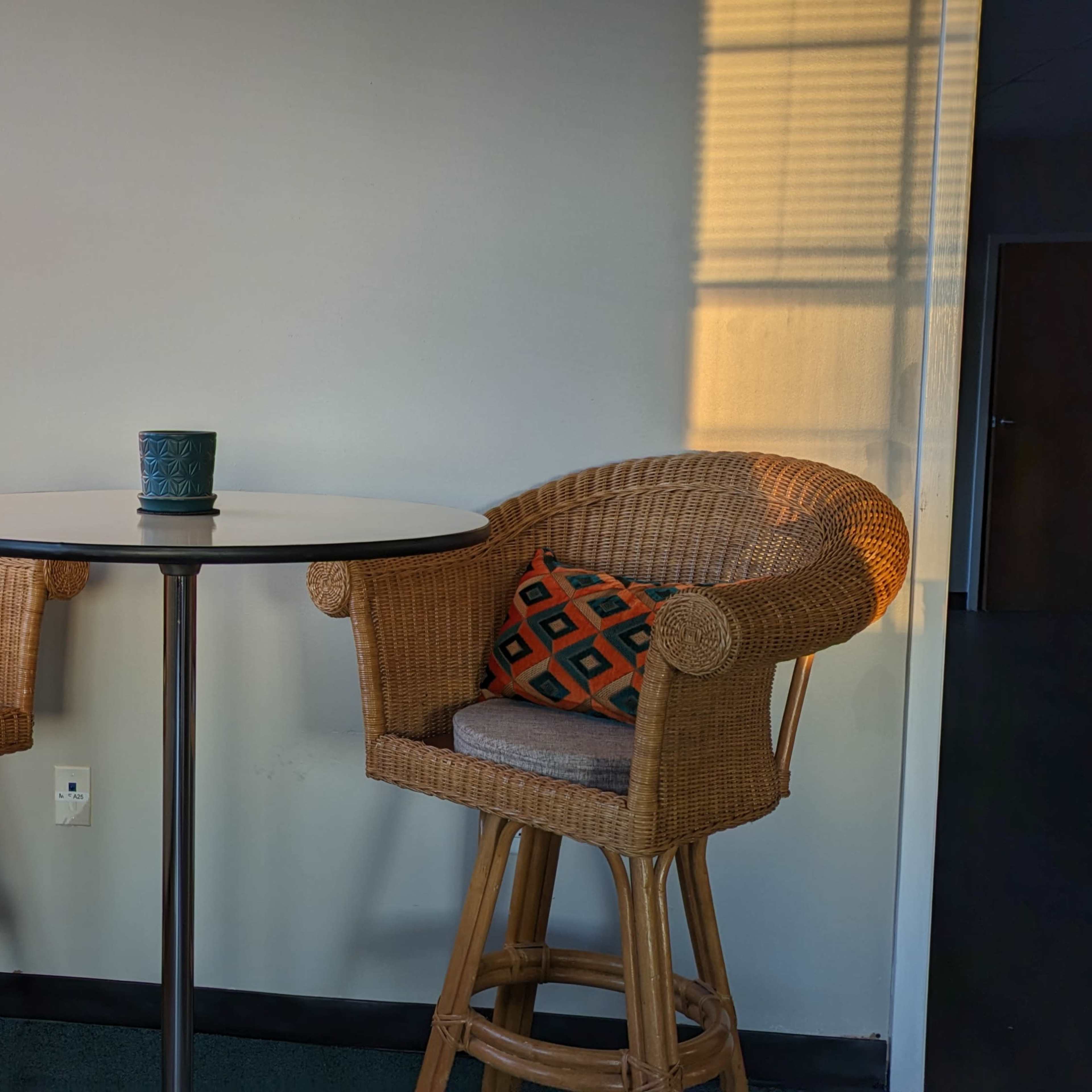 A small table with two wicker chairs, each adorned with an orange and black patterned pillow, is positioned near a window with sunlight casting shadows on the wall.