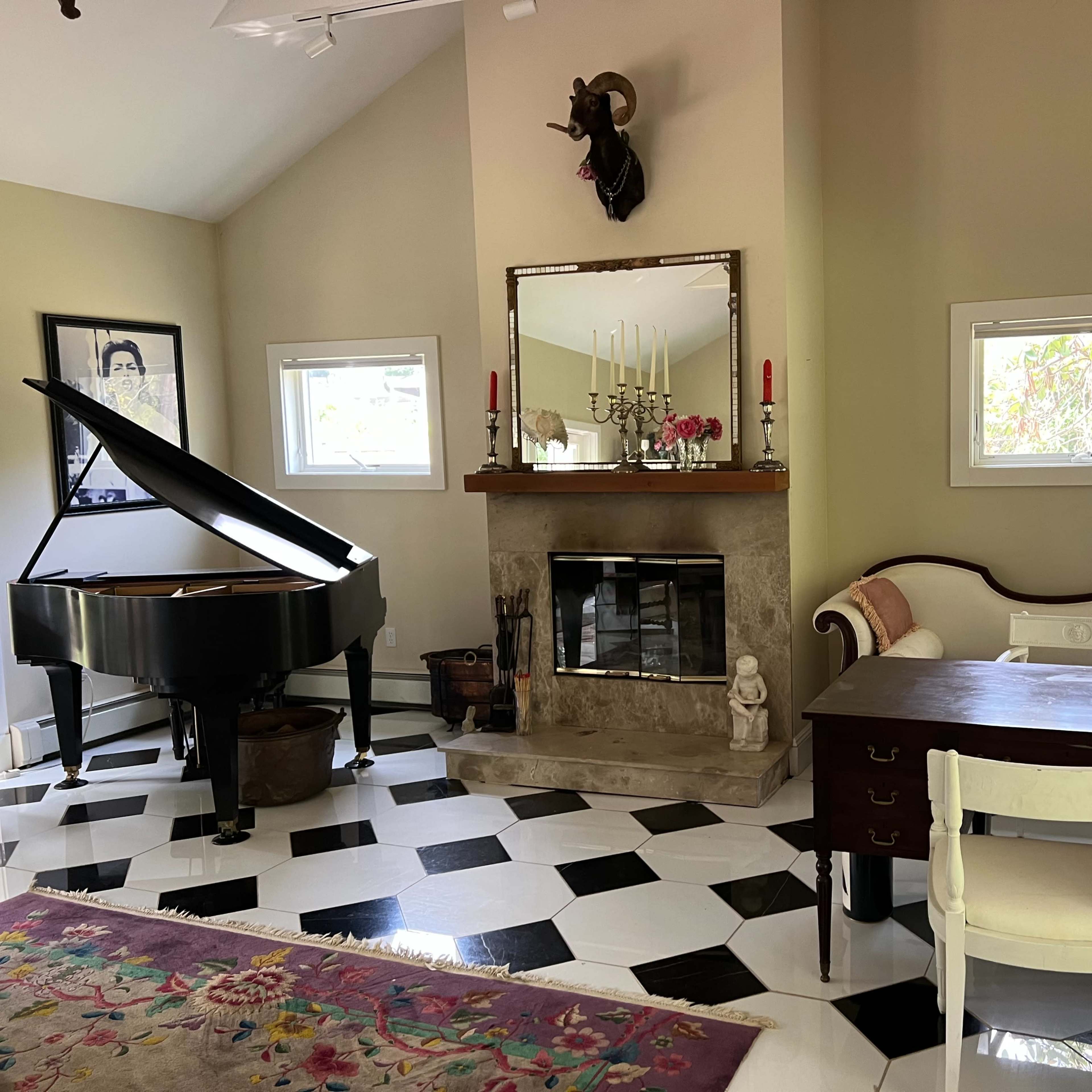 A living room featuring a grand piano, a fireplace with a mounted animal head, a vintage-style sofa, a wooden desk, and a patterned area rug on a checkered floor.
