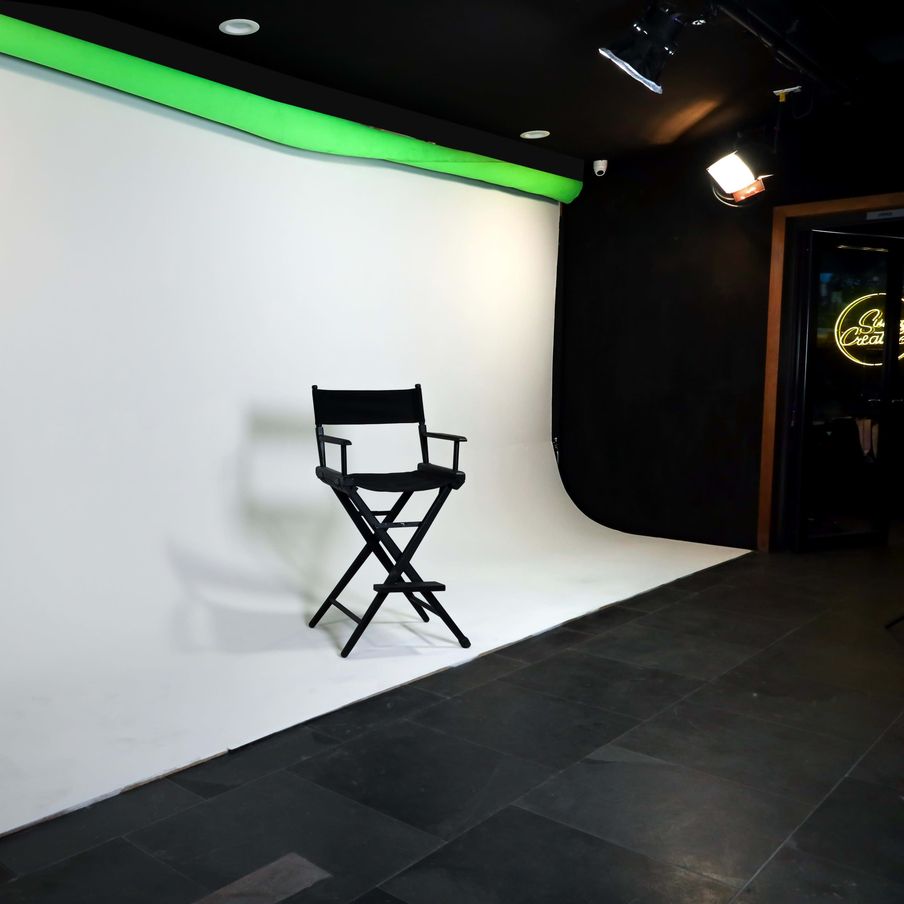 A black director's chair stands alone in front of a white backdrop with a green light strip above it.
