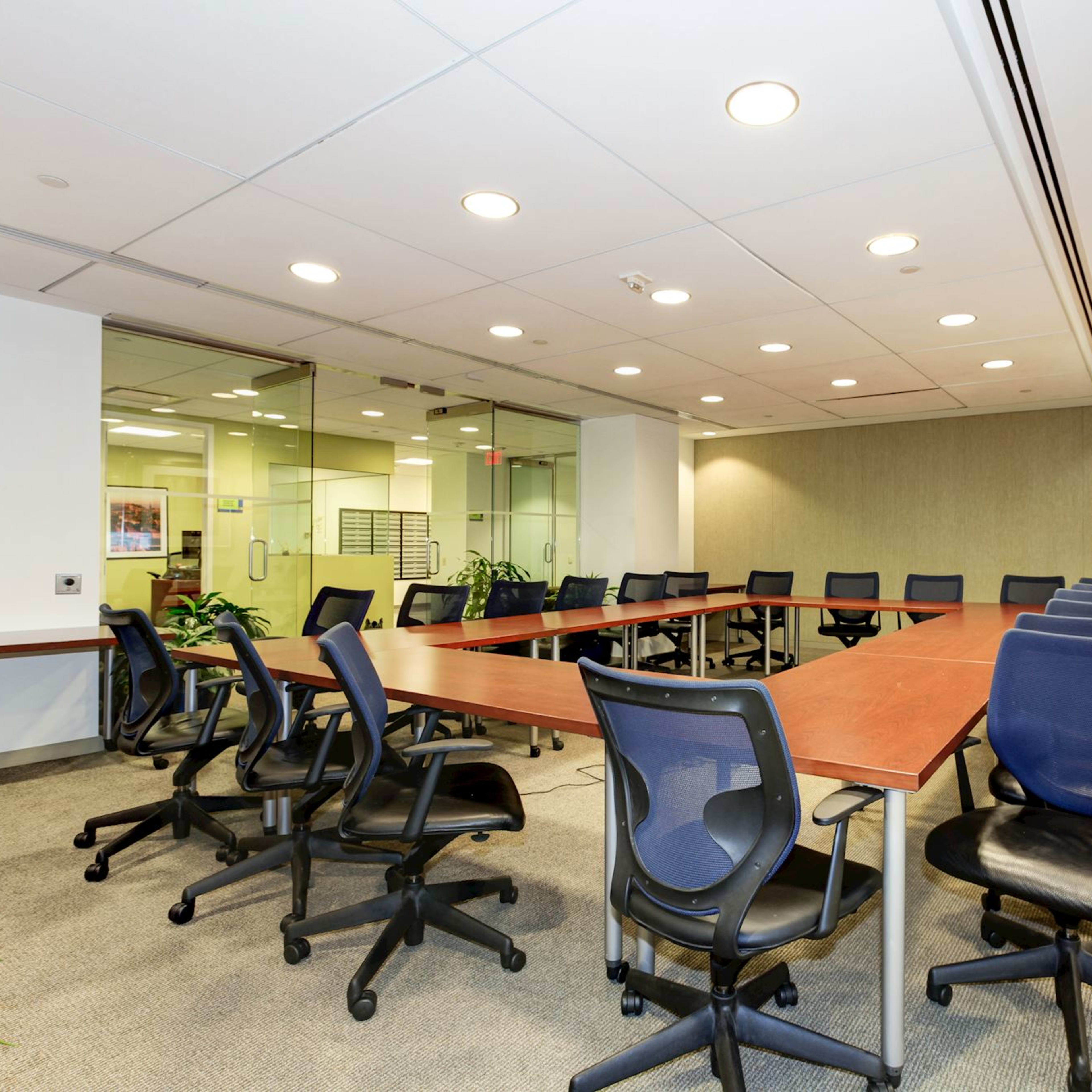 A large conference room features a rectangular table surrounded by office chairs, with glass walls and potted plants providing a view of an adjoining area.