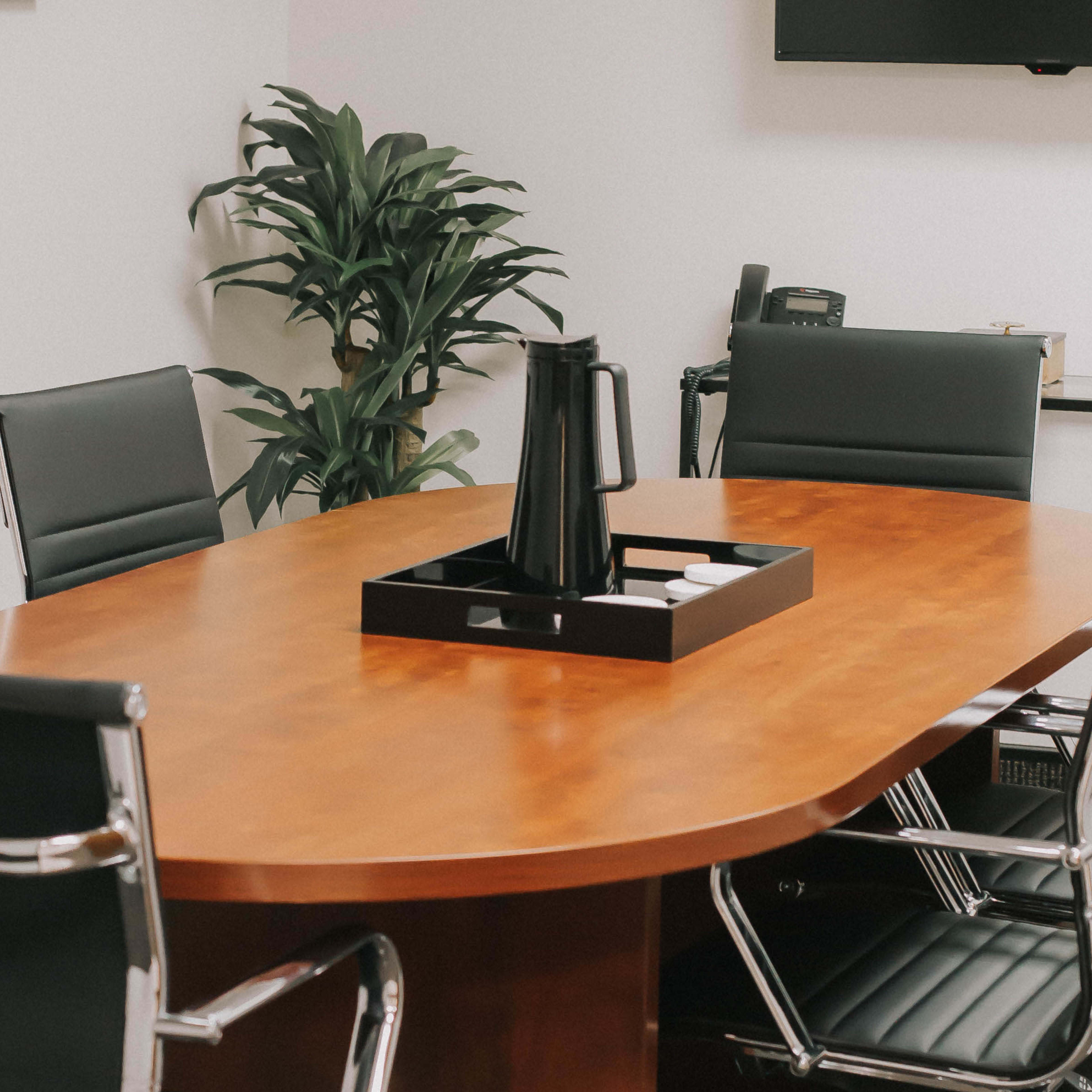 A wooden conference table surrounded by black leather chairs is set up in a modern meeting room with a plant in the corner.