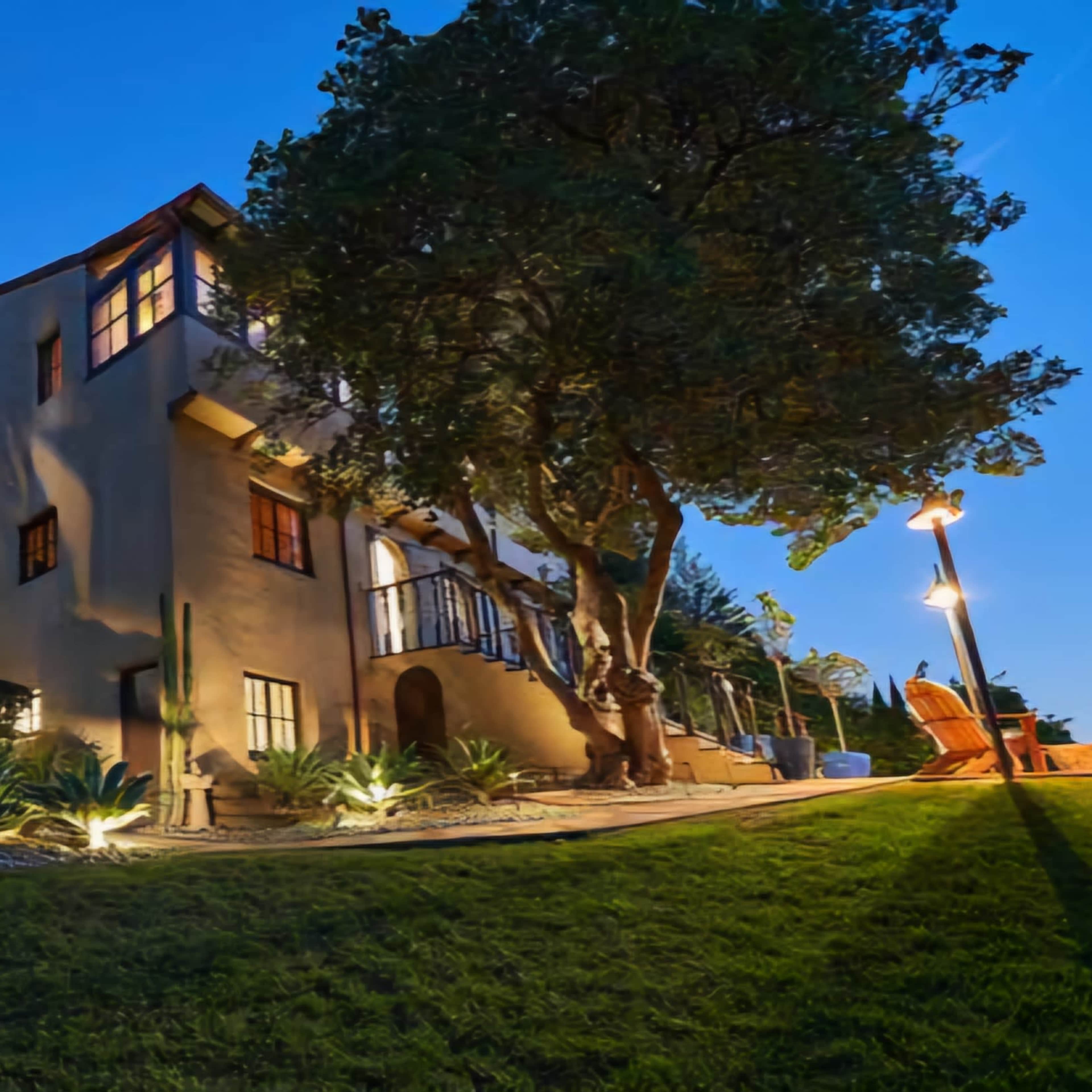 A multi-story house with large windows is set against a twilight sky, surrounded by trees and landscaped greenery.