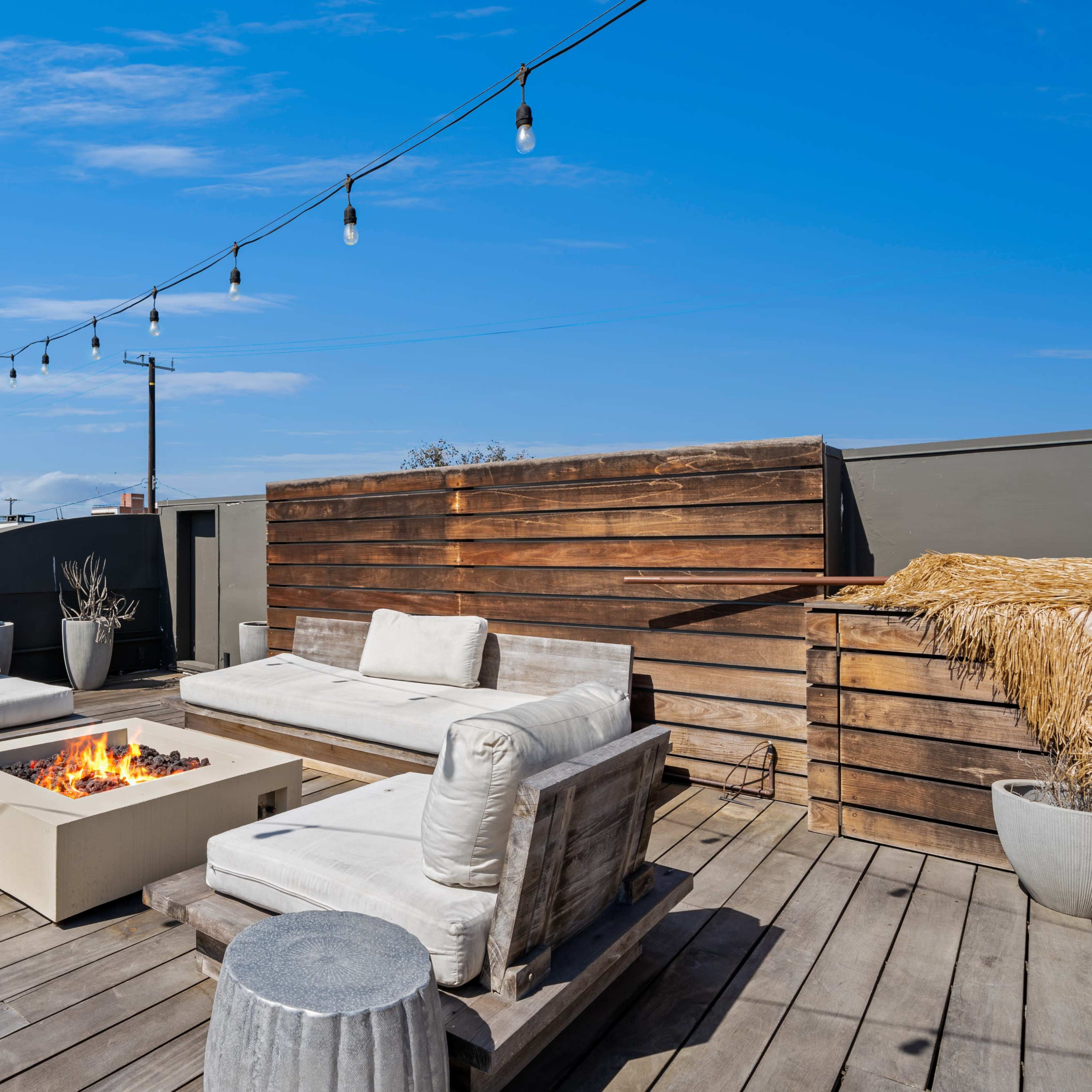 A rooftop terrace features wooden lounge furniture, a fire pit, and string lights under a clear blue sky.